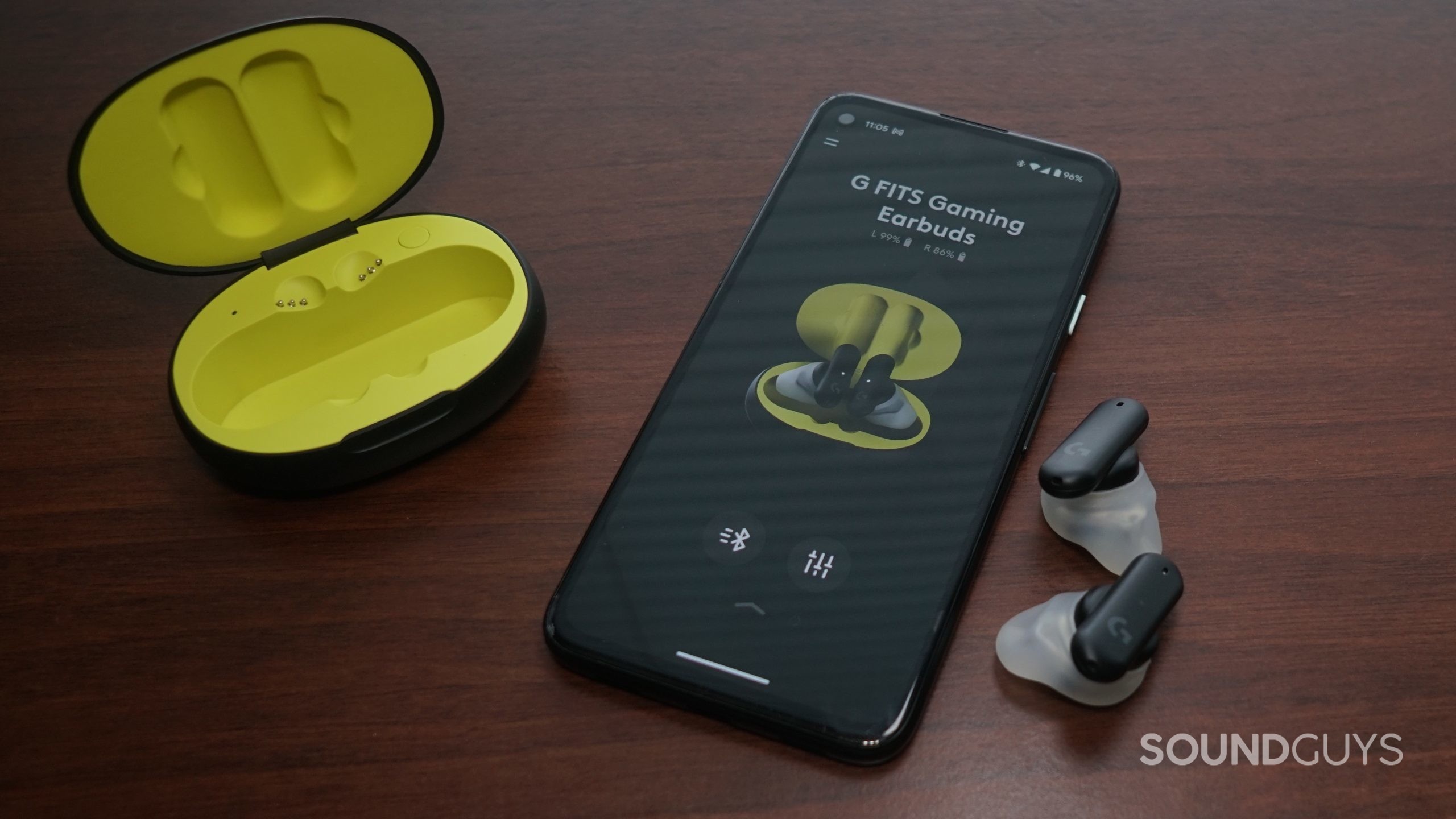 The Logitech G Fits earbuds lay on a wooden surface next to a Google Pixel 4a running the Logitech G Fits app.