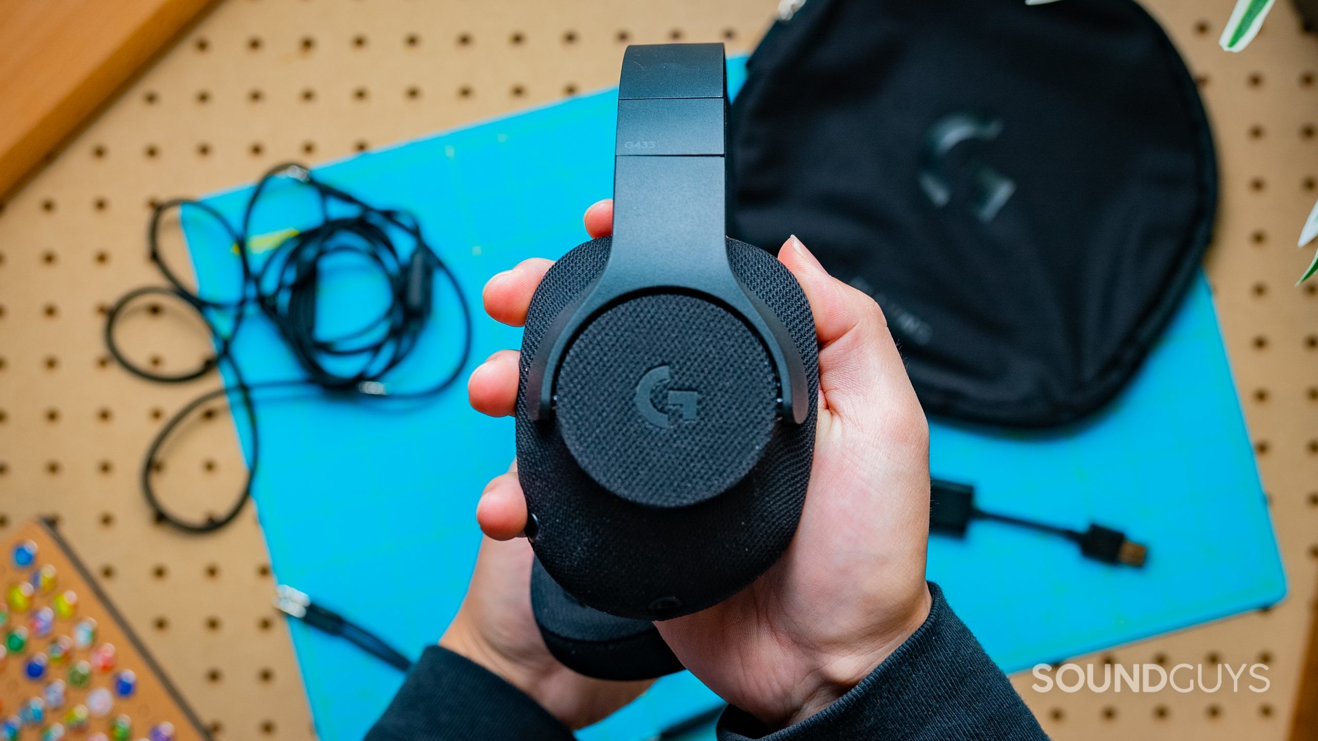 The Logitech G433 being held in someone's hands with its accessories on the table behind it.