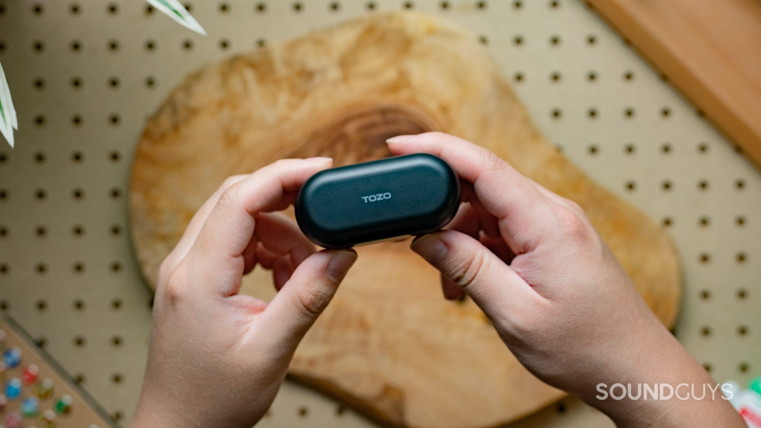 The TOZO T12 charging case being held up by two hands over a table.