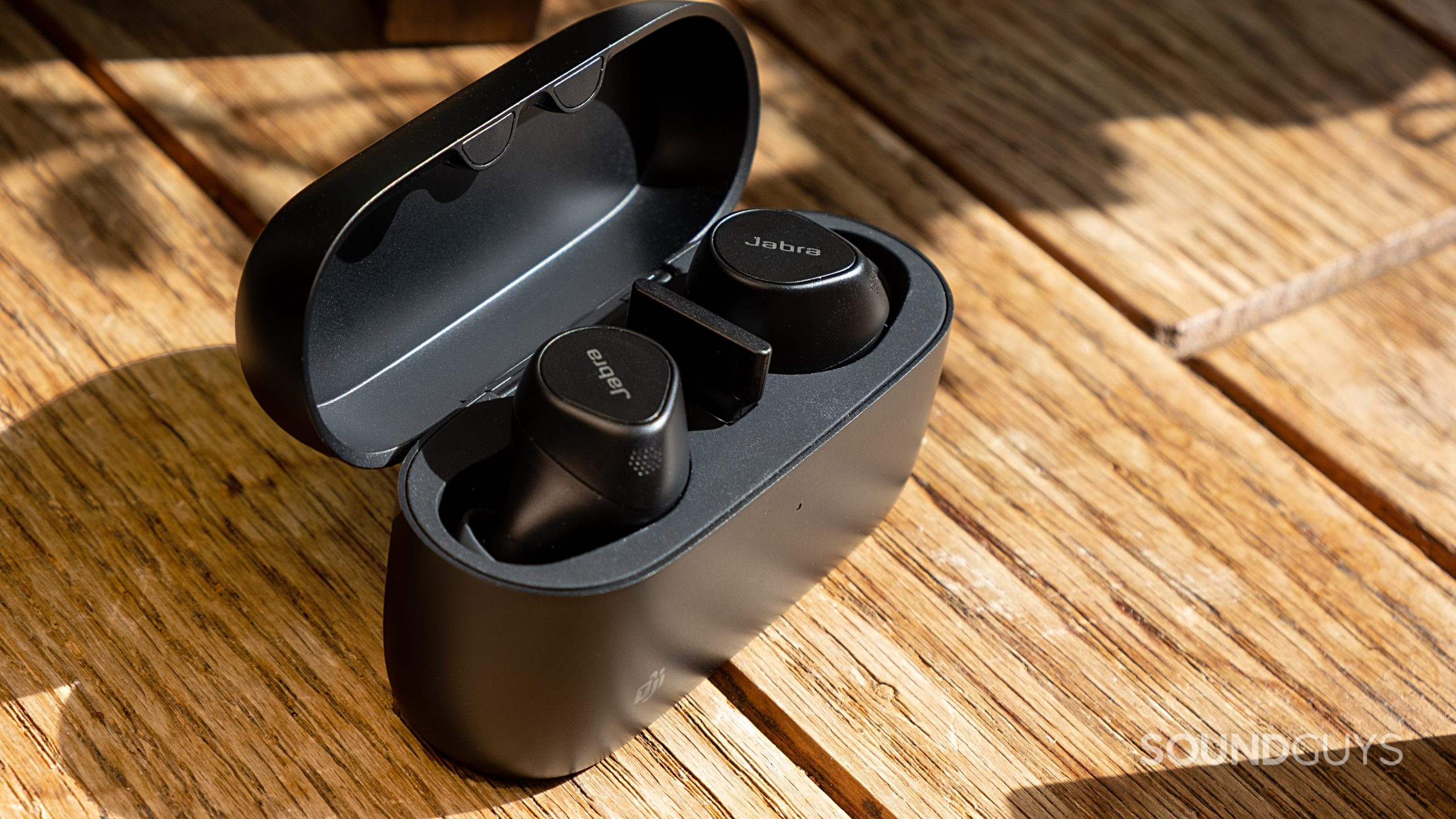 On a wood surface the Jabra Evolve2 Buds are seated in the case with the lid open showing the buds and dongle.
