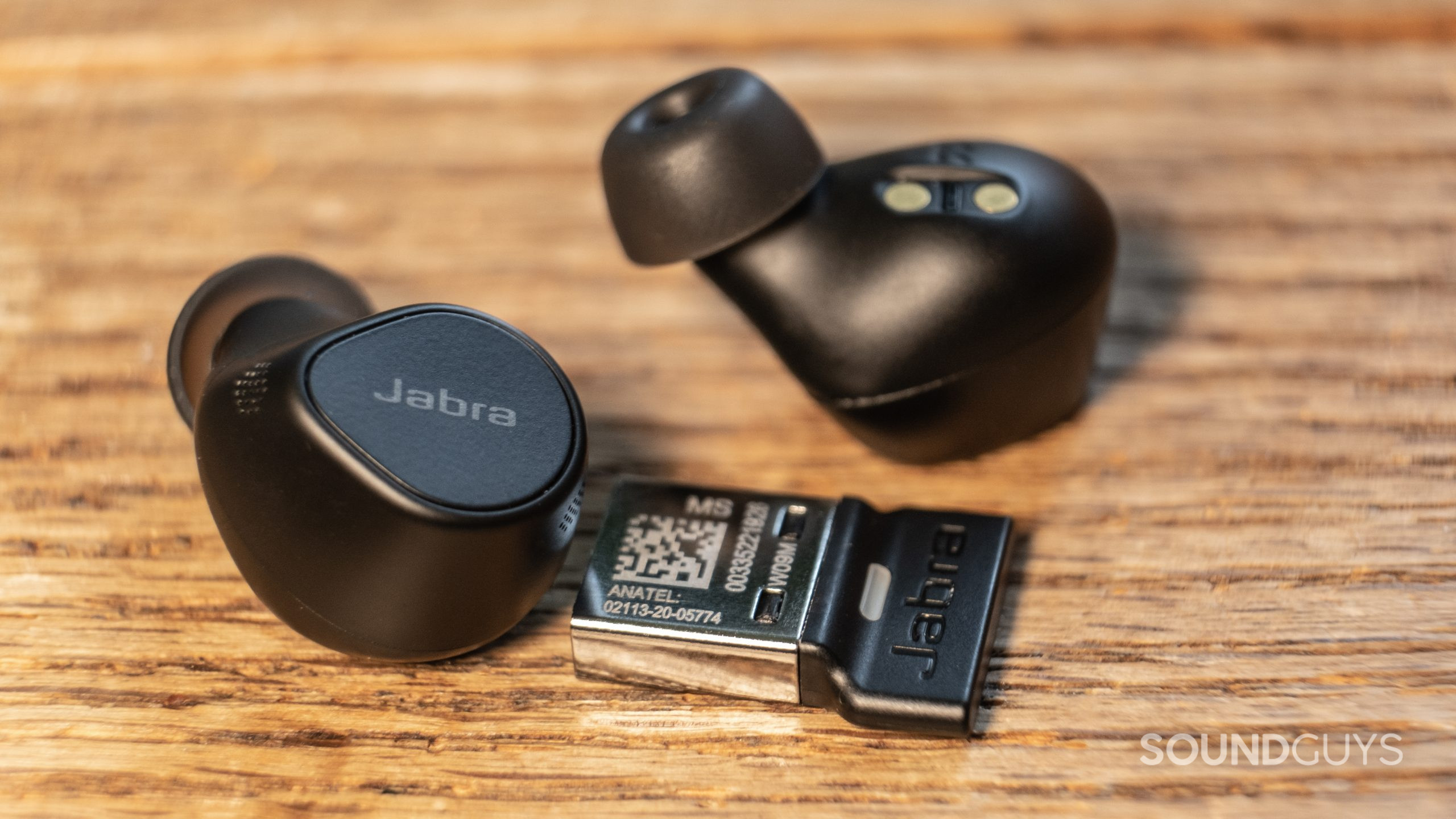 A close up shows the Jabra Evolve2 Buds and dongle out of the case on a wood surface.