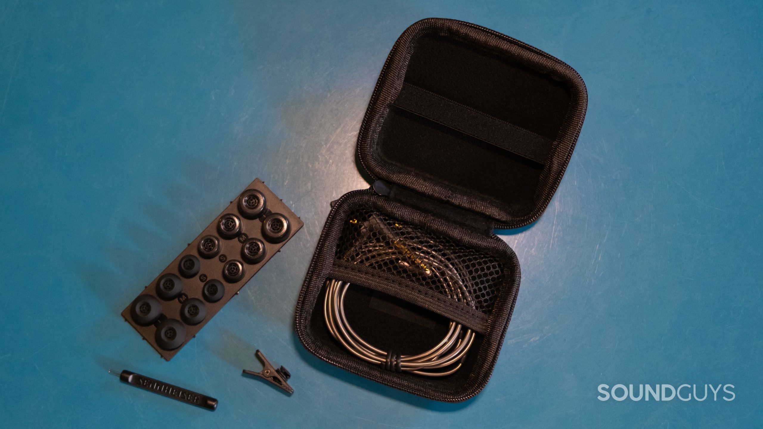 On a turquoise surface are the opened case, cleaning tool, lapel clip, extra cable, and tray of ear tips with the Sennheiser IE 600.