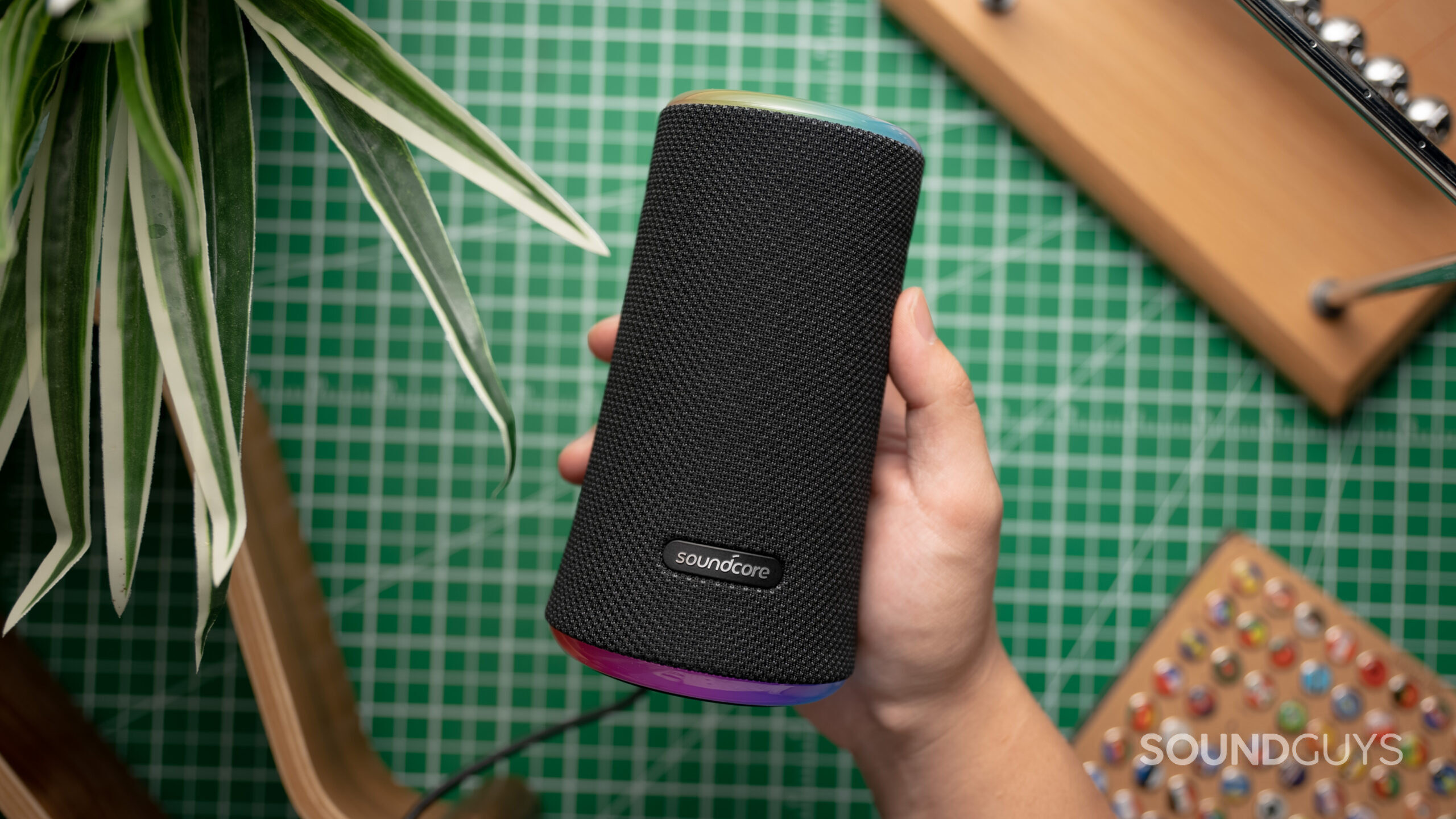 Anker Soundcore Flare 2 being held up by a hand against a flat surface with a plant and wooden objects in the background.