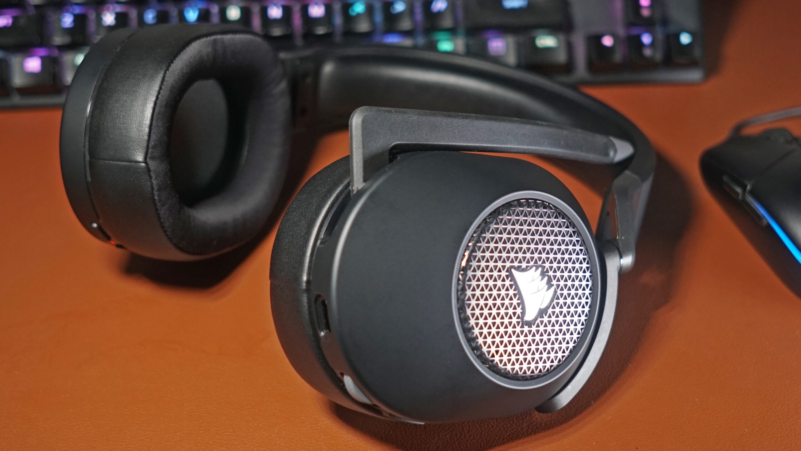 The Corsair HS65 Wireless gaming headset lays on a leather surface next to a Logitech gaming mouse and in front of a HyperX mechanical gaming keyboard.