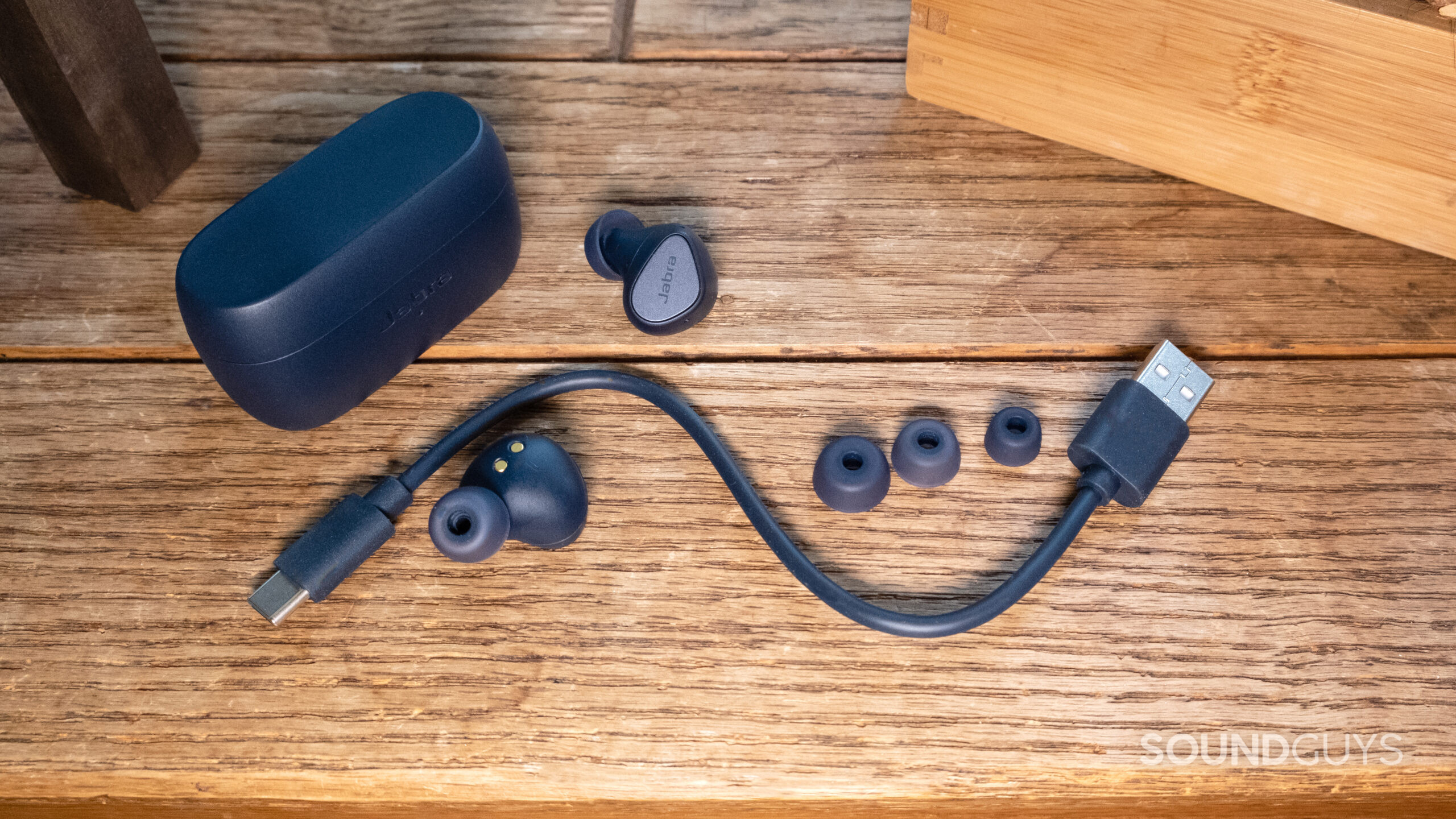 A top down shot shows the closed case, earbuds from different angles, three ear tip sizes, and charge cable for the Jabra Elite 4 on a wood surface.