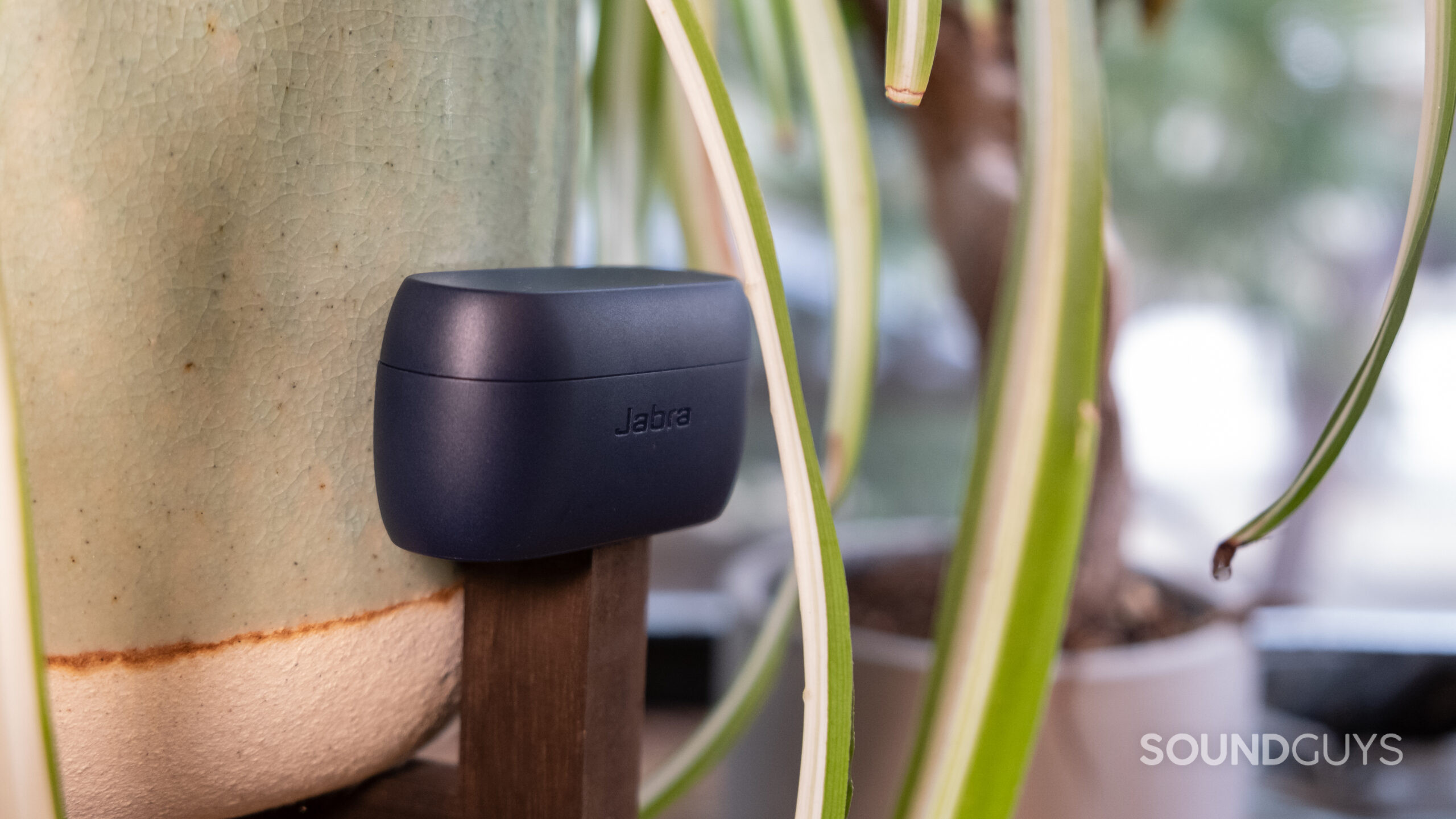 The closed battery charging case of the Jabra Elite 4 sits on a wood stand beside a ceramic pot with fern leaves around it.