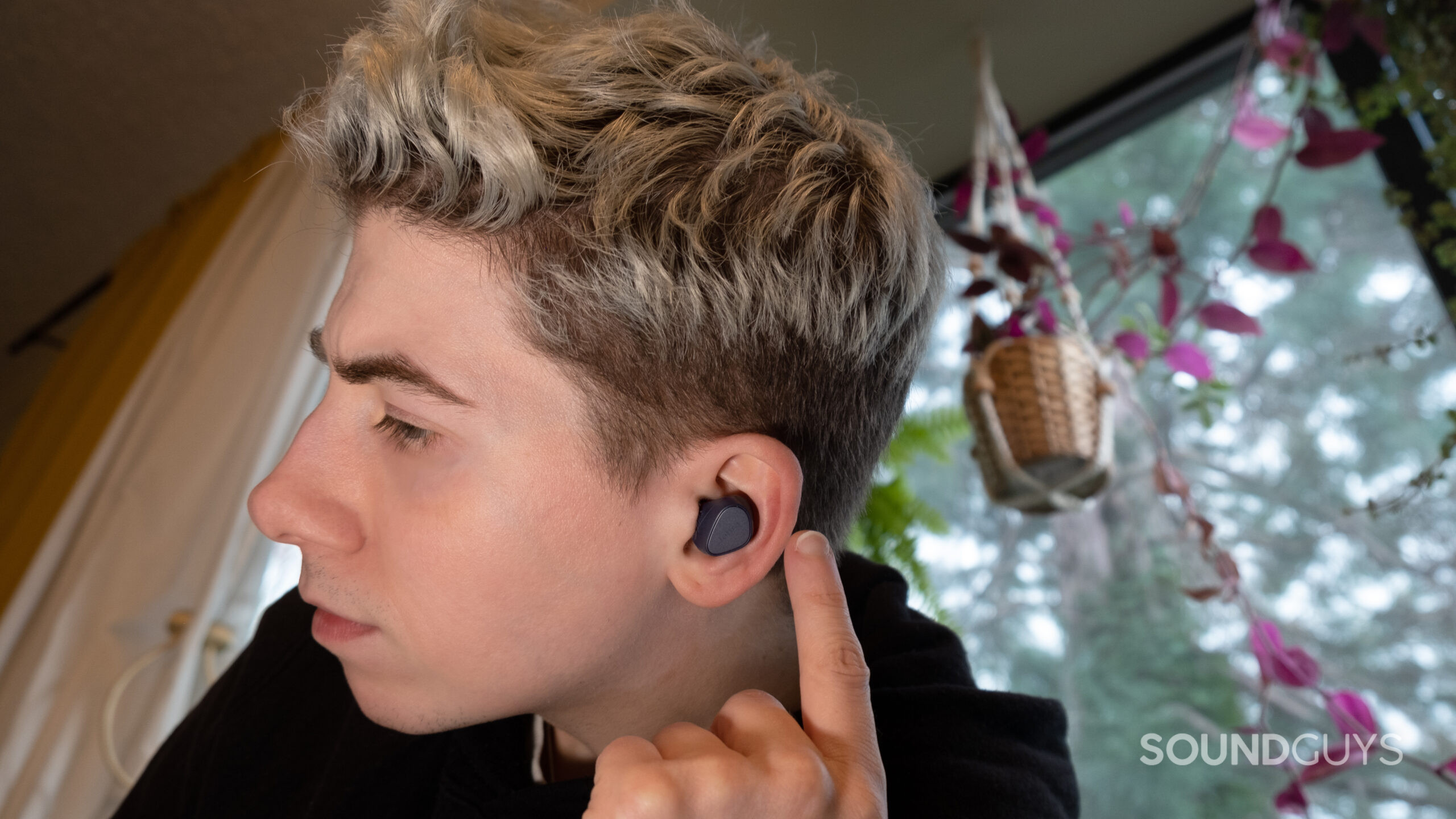 A man faces left about to press the button on the Jabra Elite 4 with a window and hanging plants in the background.