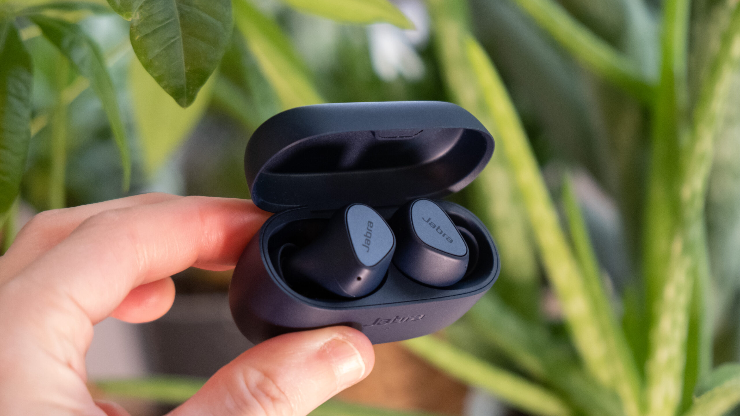 A hand holds the Jabra Elite 4 open in front of green foliage.