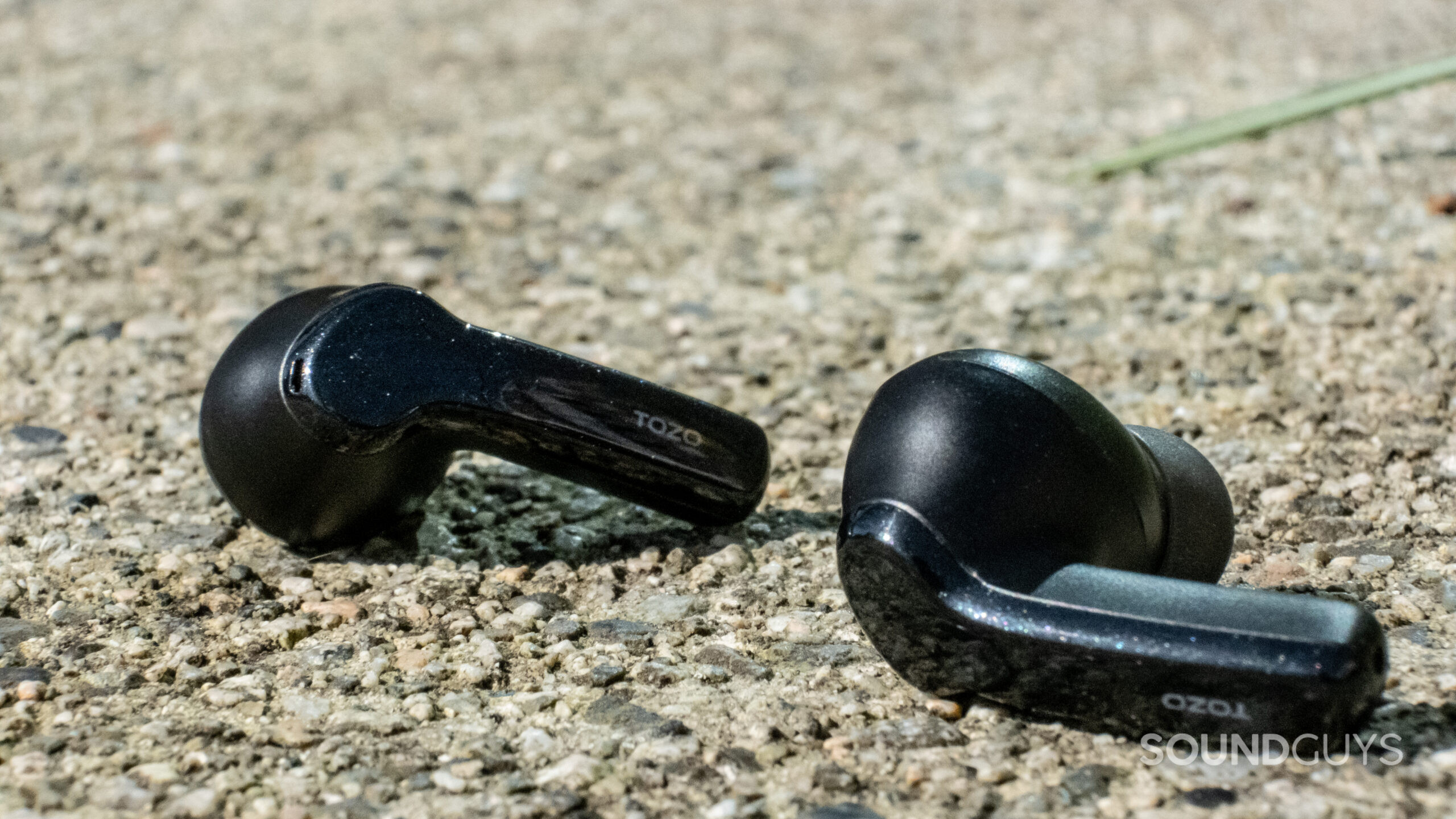 A macro shot of the TOZO NC2 buds shows the different textures and shades of black in sunlight on concrete.