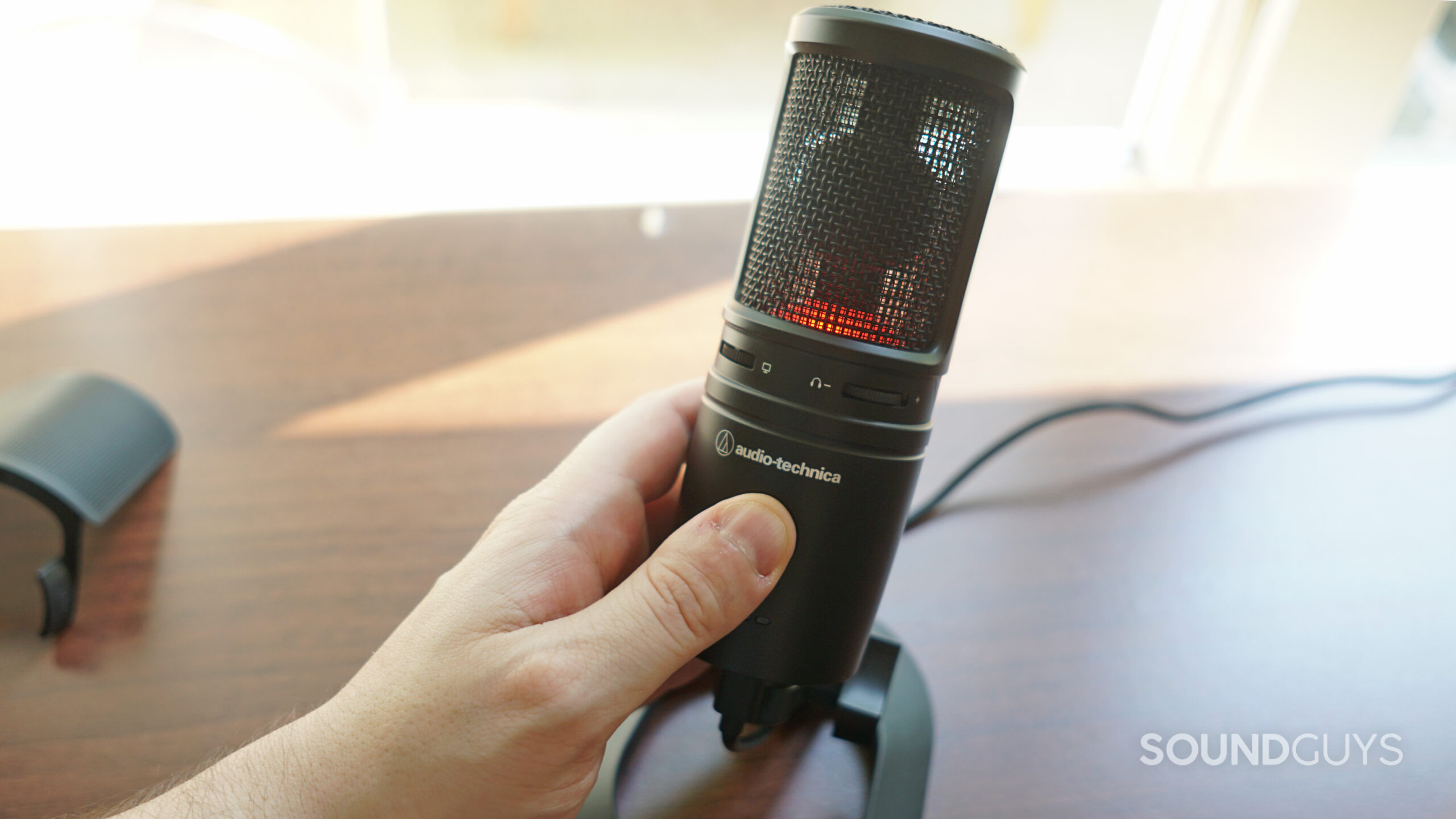 The AT2020USB-XP on top of a wooden surface with a hand around it, the red LED is lit up indicating that it's muted.