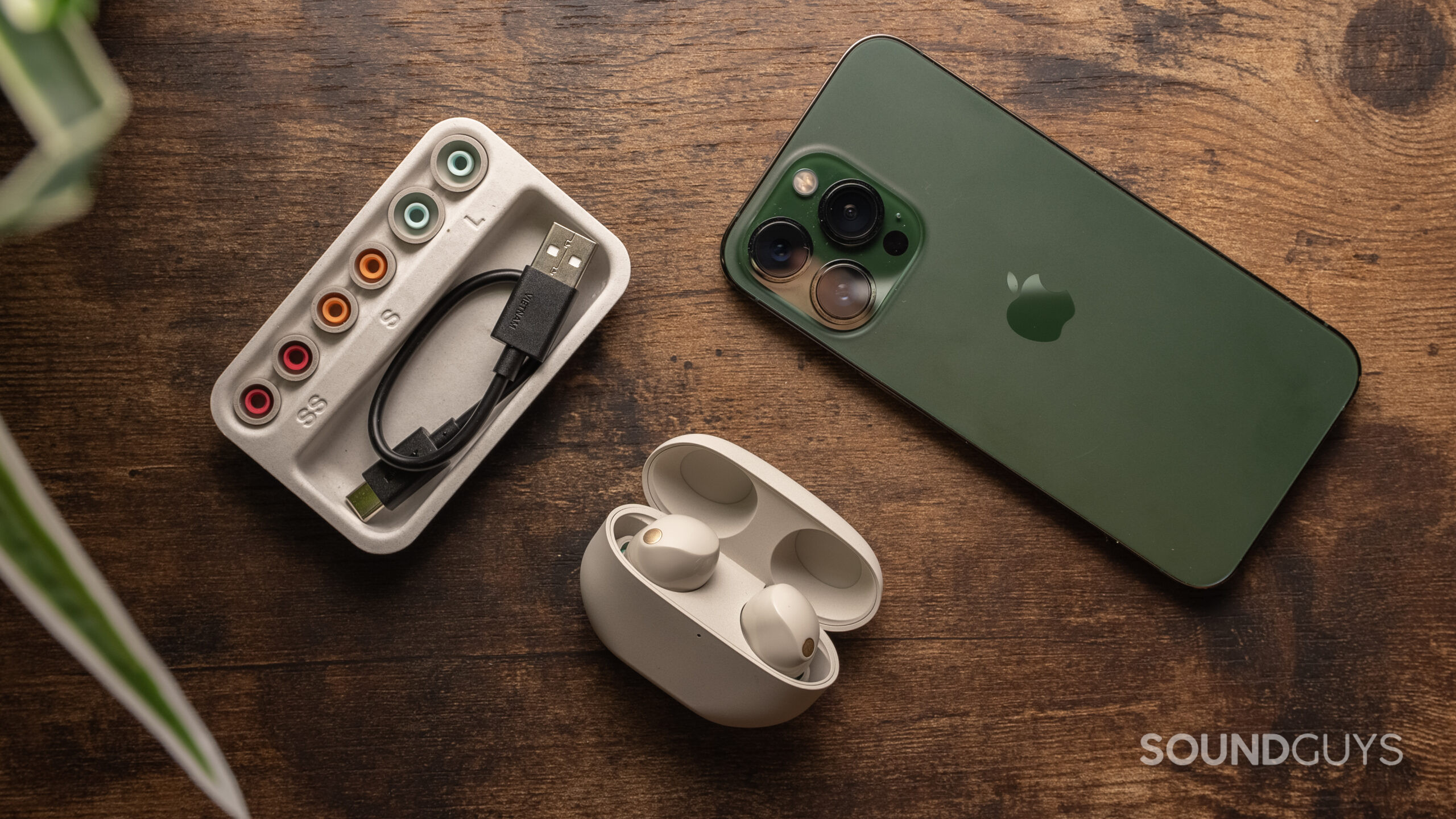 The Sony WF-1000XM5 charging case open with the earbuds inside beside a green iPhone 13 Pro and the ear tip selection and USB charging cable