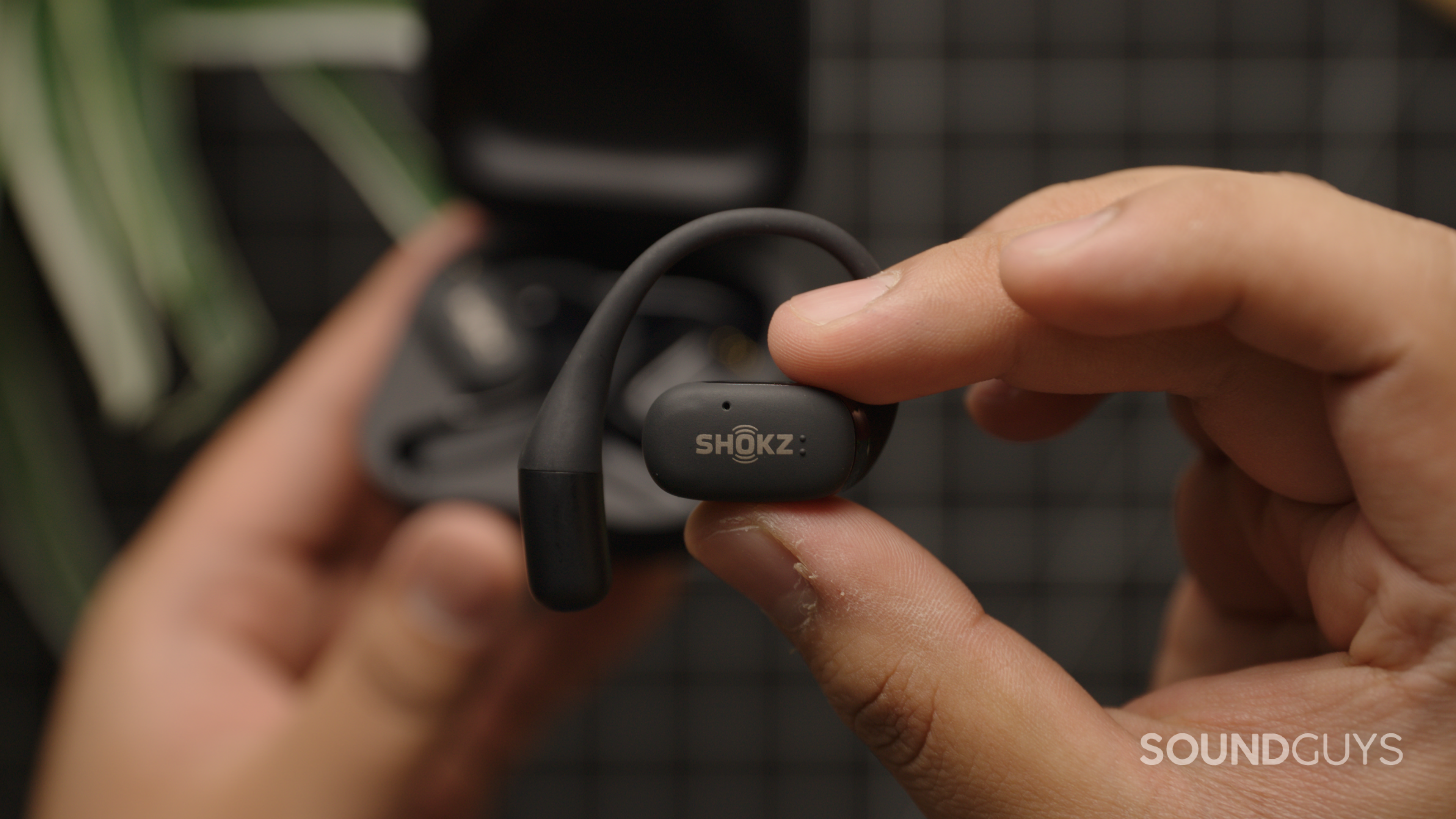 A hand holds the Shokz OpenFit with the case out of focus in the background.