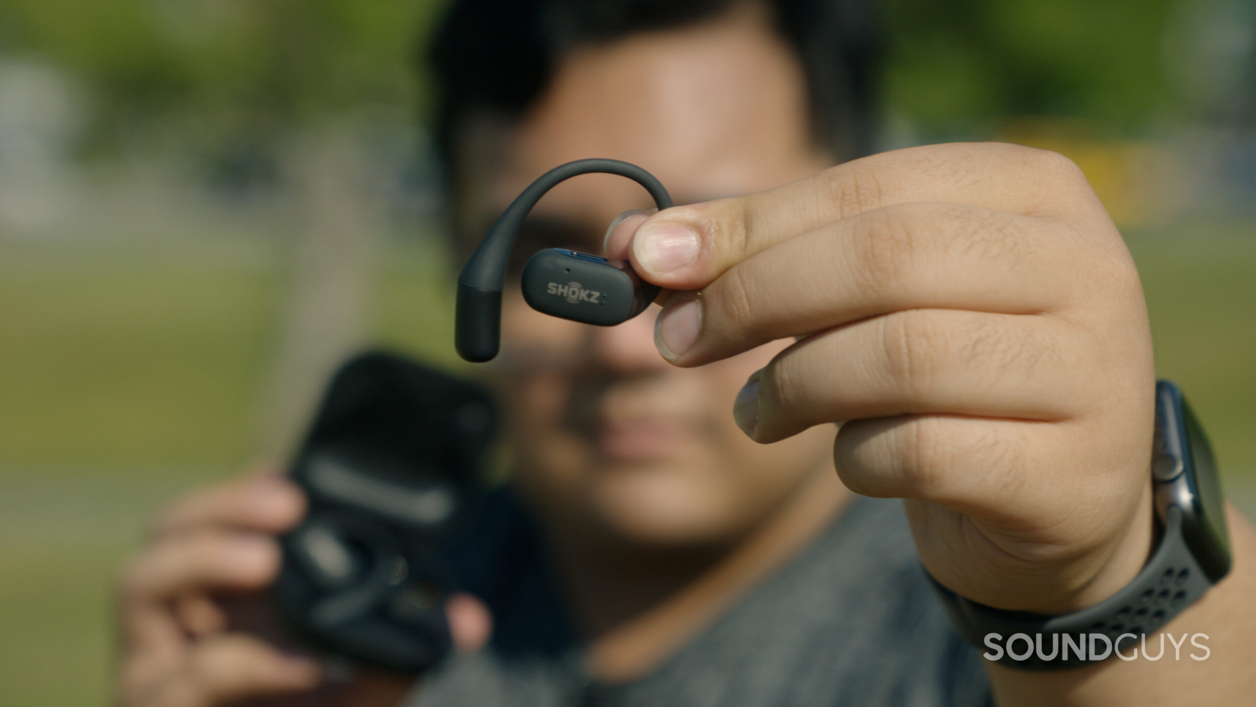 A man out of focus holds the Shokz OpenFit in focus.
