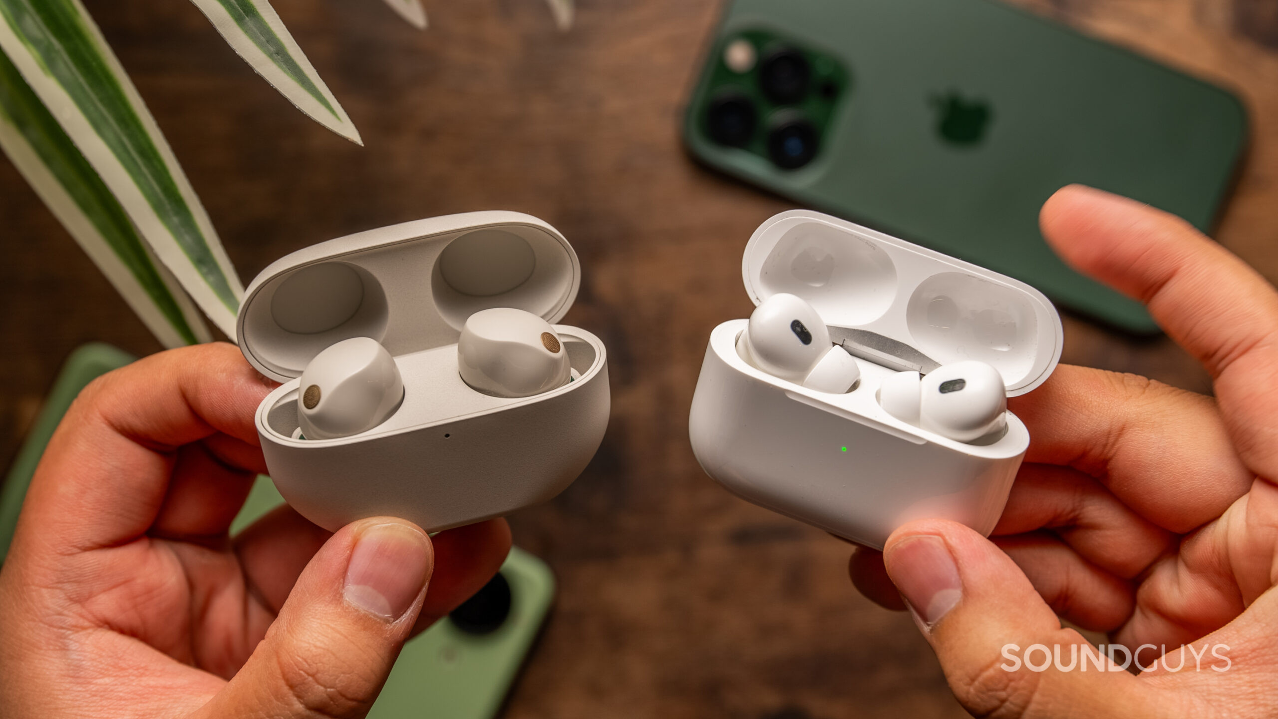 A hand holds the Sony WF-1000XM5 and Apple AirPods Pro 2 cases in each hand.