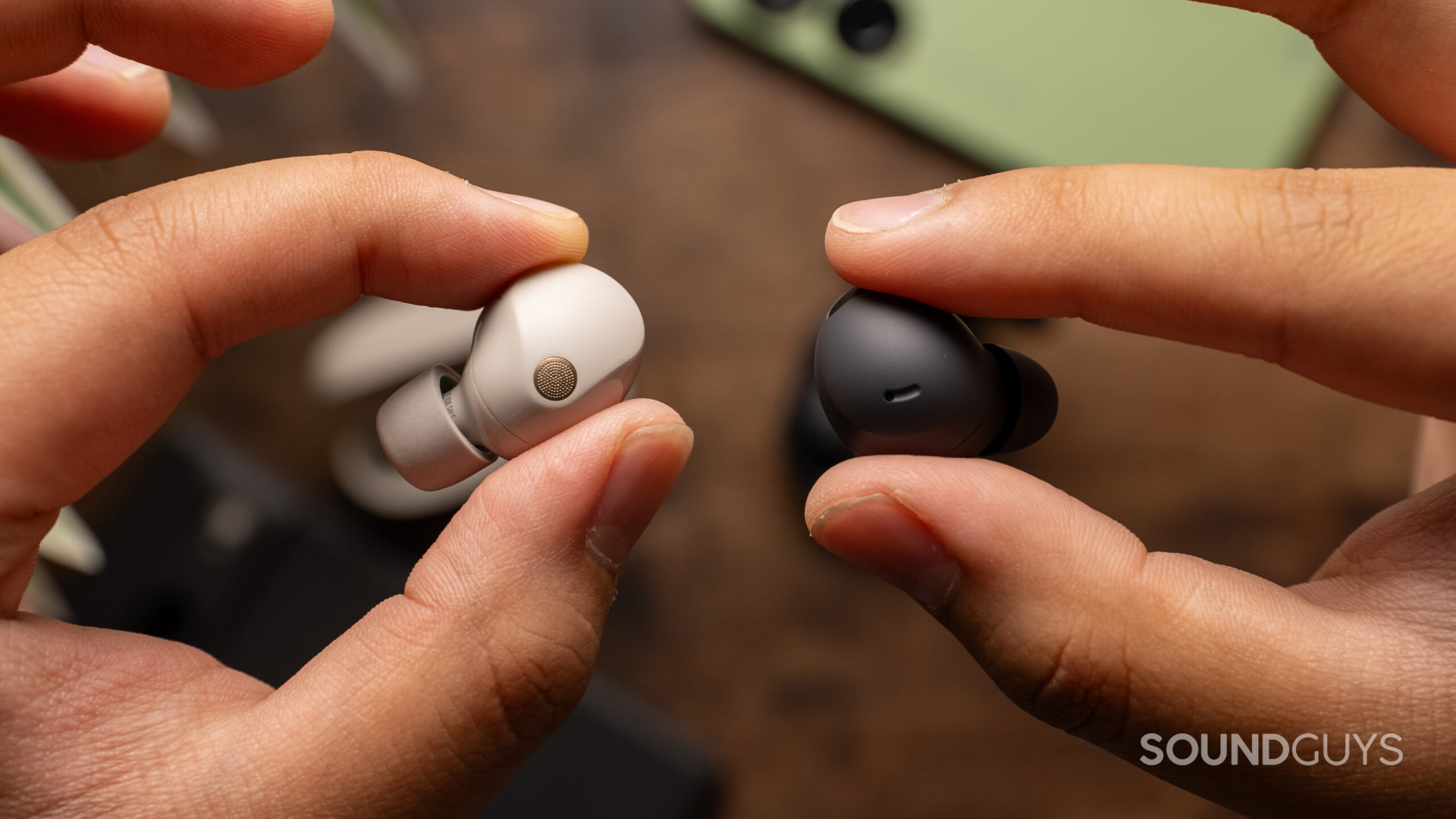 Two hands hold one of each Sony WF-1000XM5 and Samsung Galaxy Buds 2 Pro above a wood table.