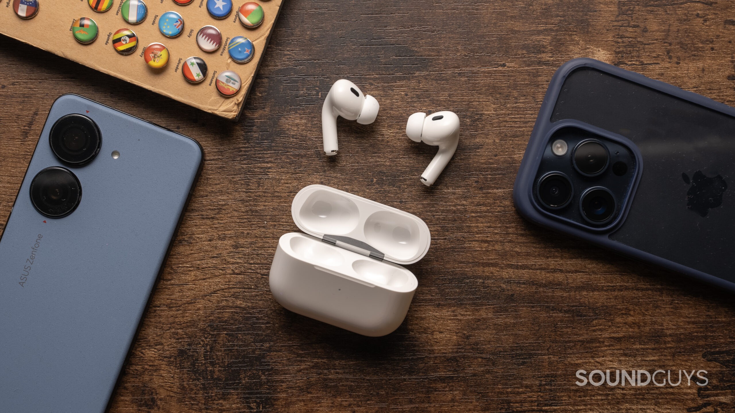 Apple AirPods Pro (2nd generation) resting on a dark wood tabletop with the lid open and buds on display surrounded by phones.