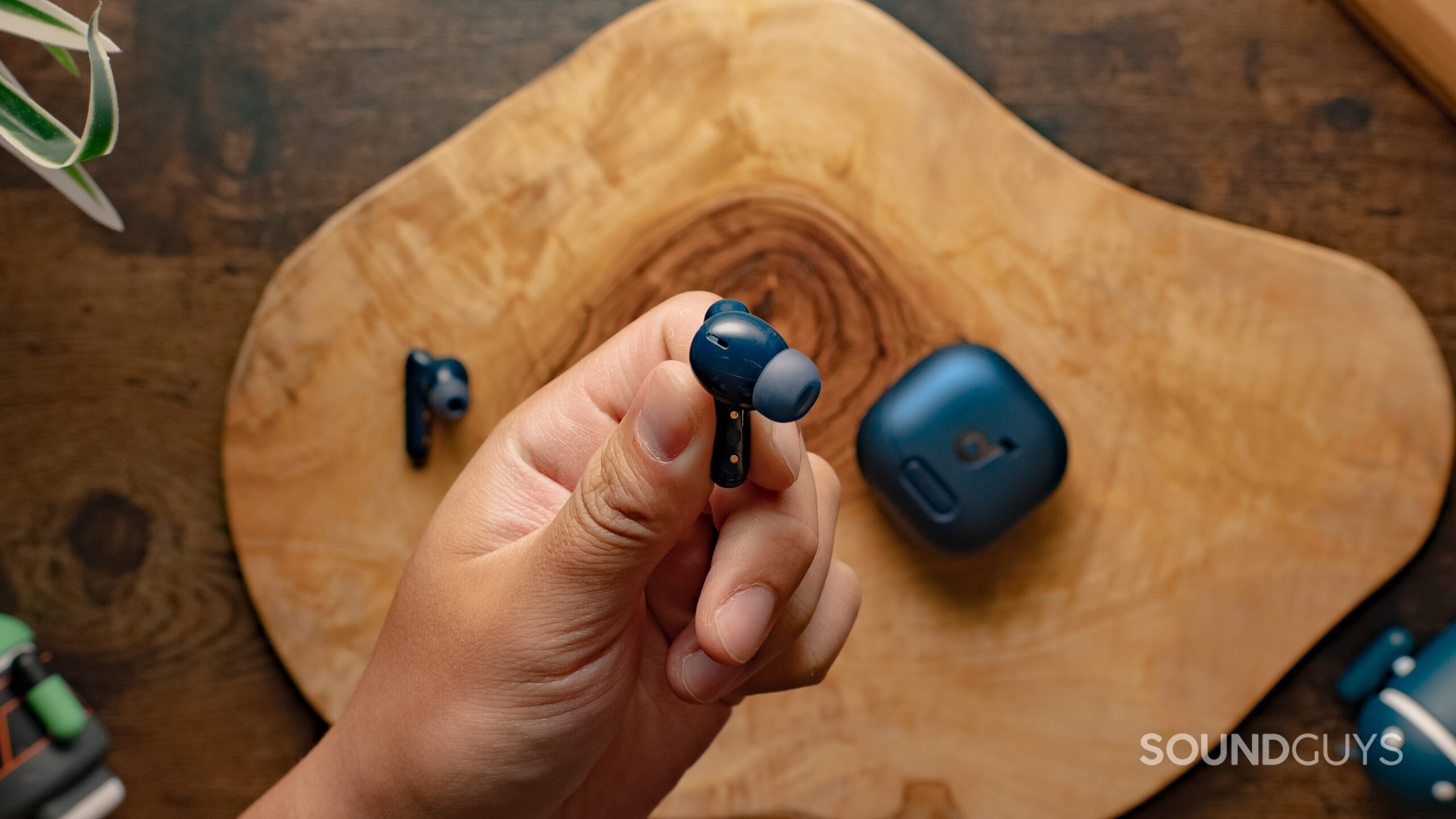 A hand holds one of the Anker Soundcore Liberty 4 NC by the stem with the case on a wood surface in the background.