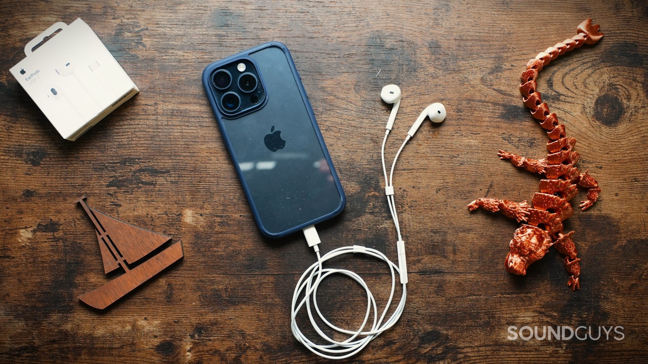 Apple EarPods (USB-C) plugged into a blue iPhone on a wooden table next to a copper 3D printed Dragon and a wooden cutout of a boat.