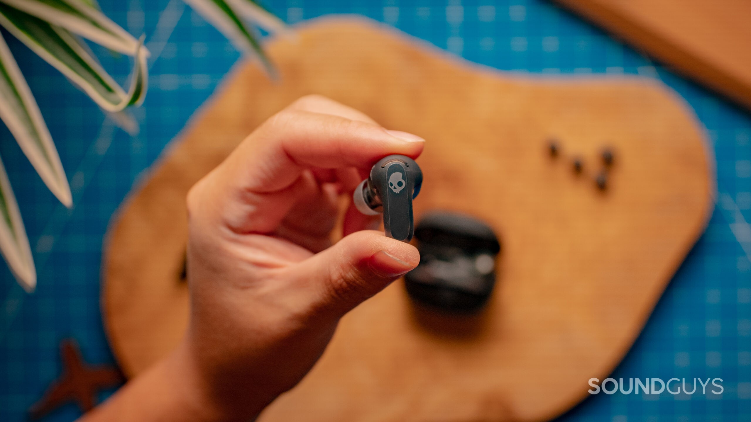 A hand holds up the Skullcandy Rail ANC showing the outer side with a wood surface in the background.