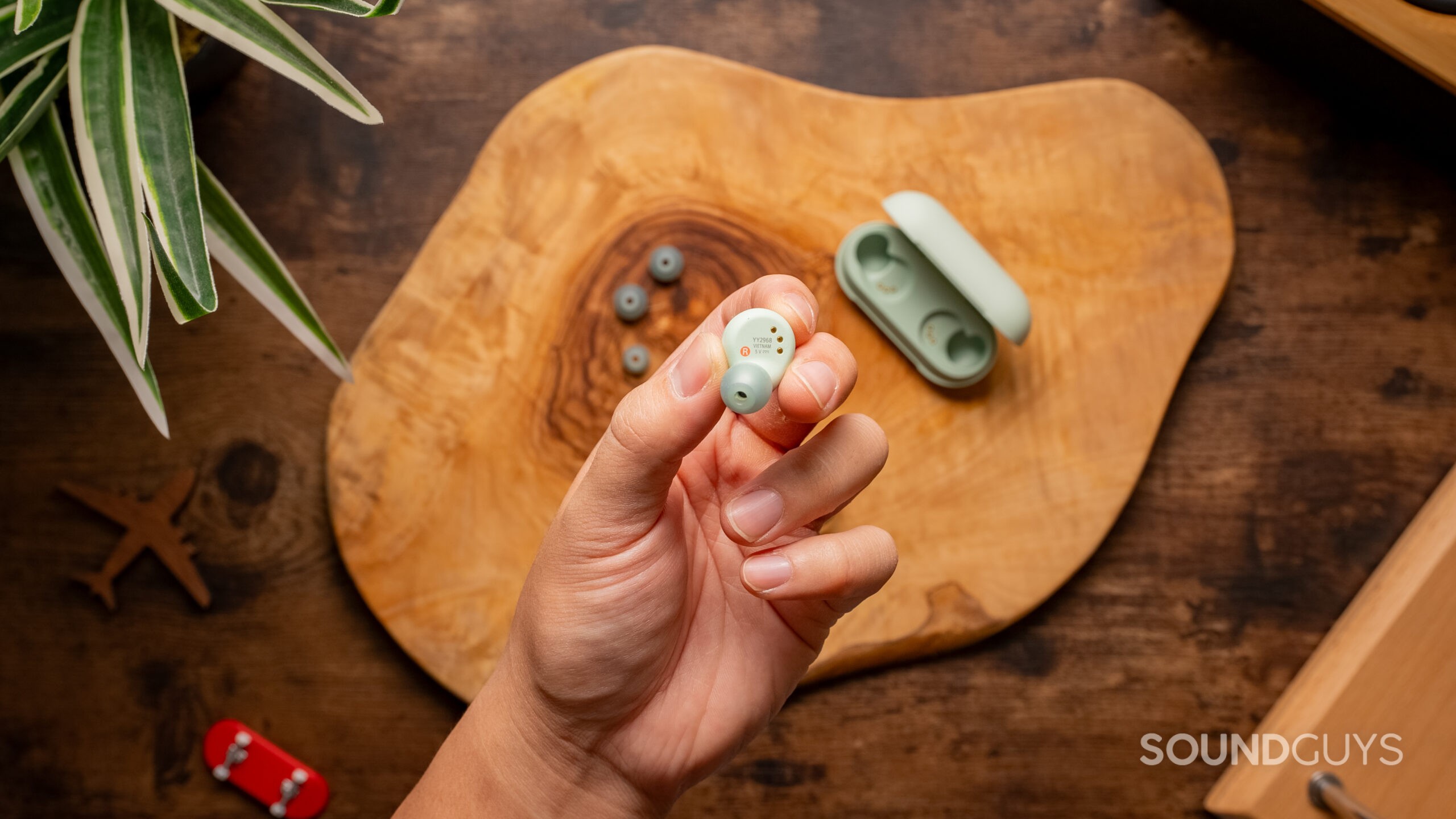 A hand holds one of the Sony WF-C700N earbuds with the open case in the background on a wood table.