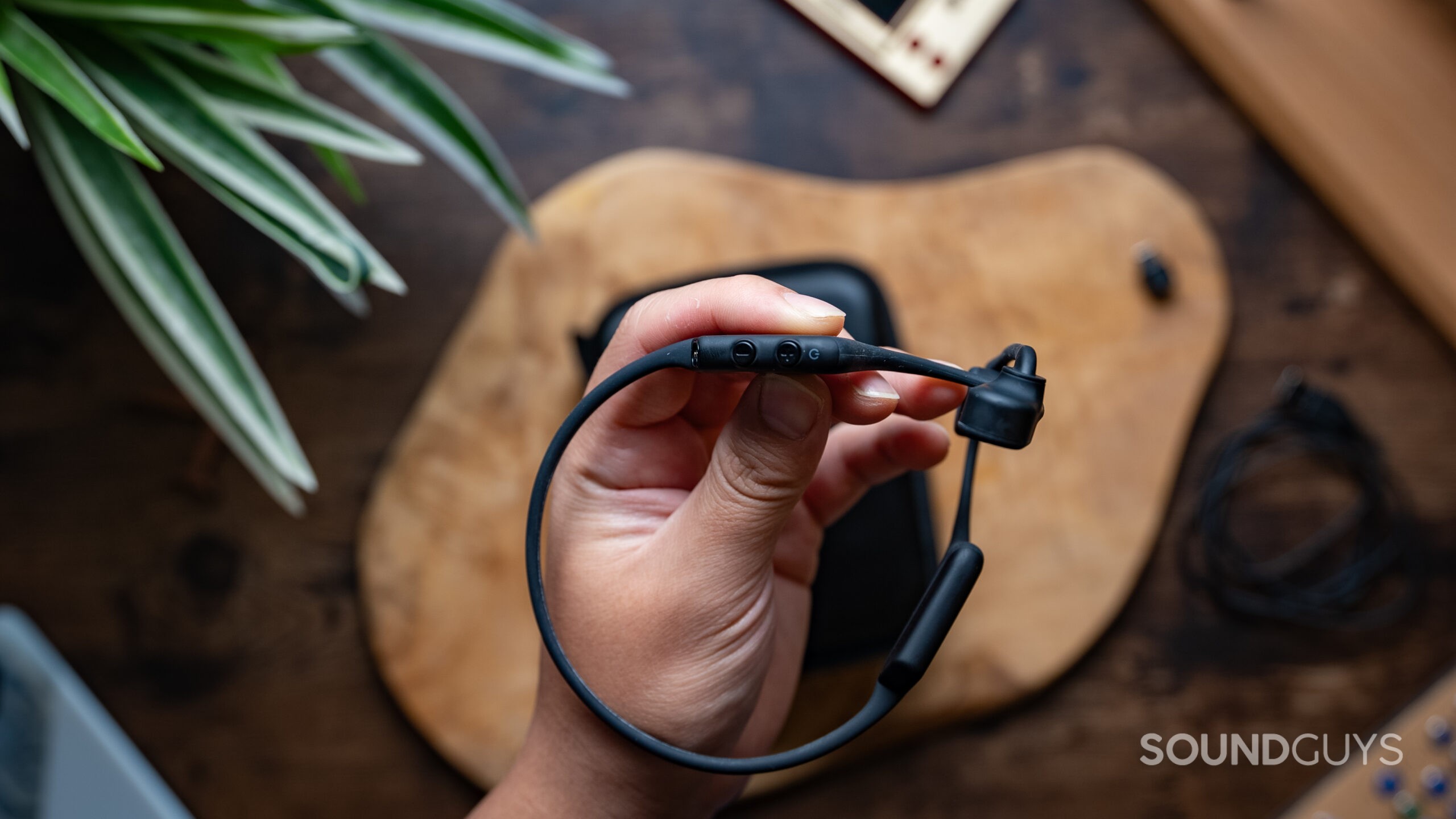 A hand holds the Shokz OpenComm2 UC headset with a blurred wood background.