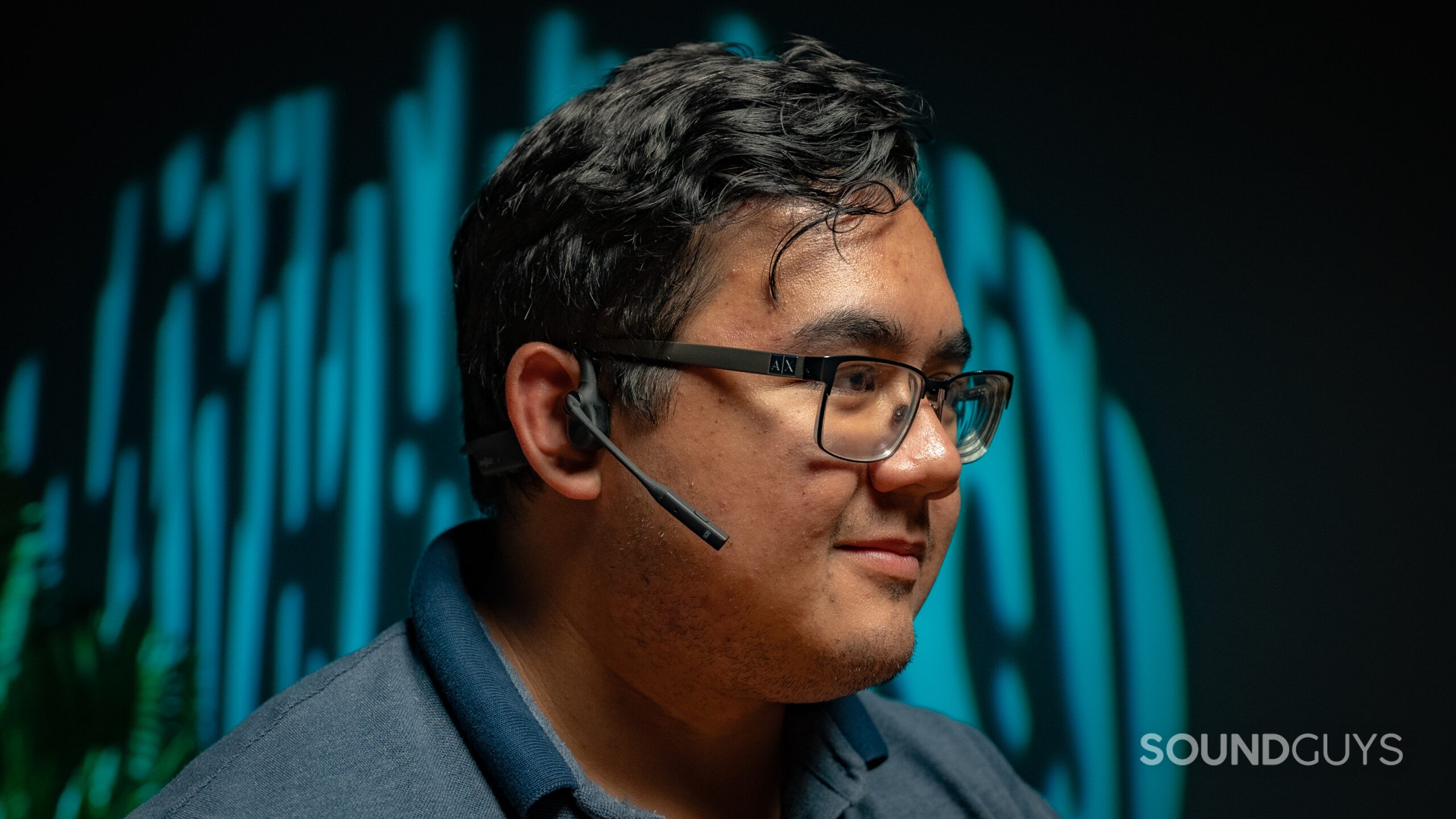 A man facing right wears the Shokz OpenComm2 UC with a green and black backdrop.