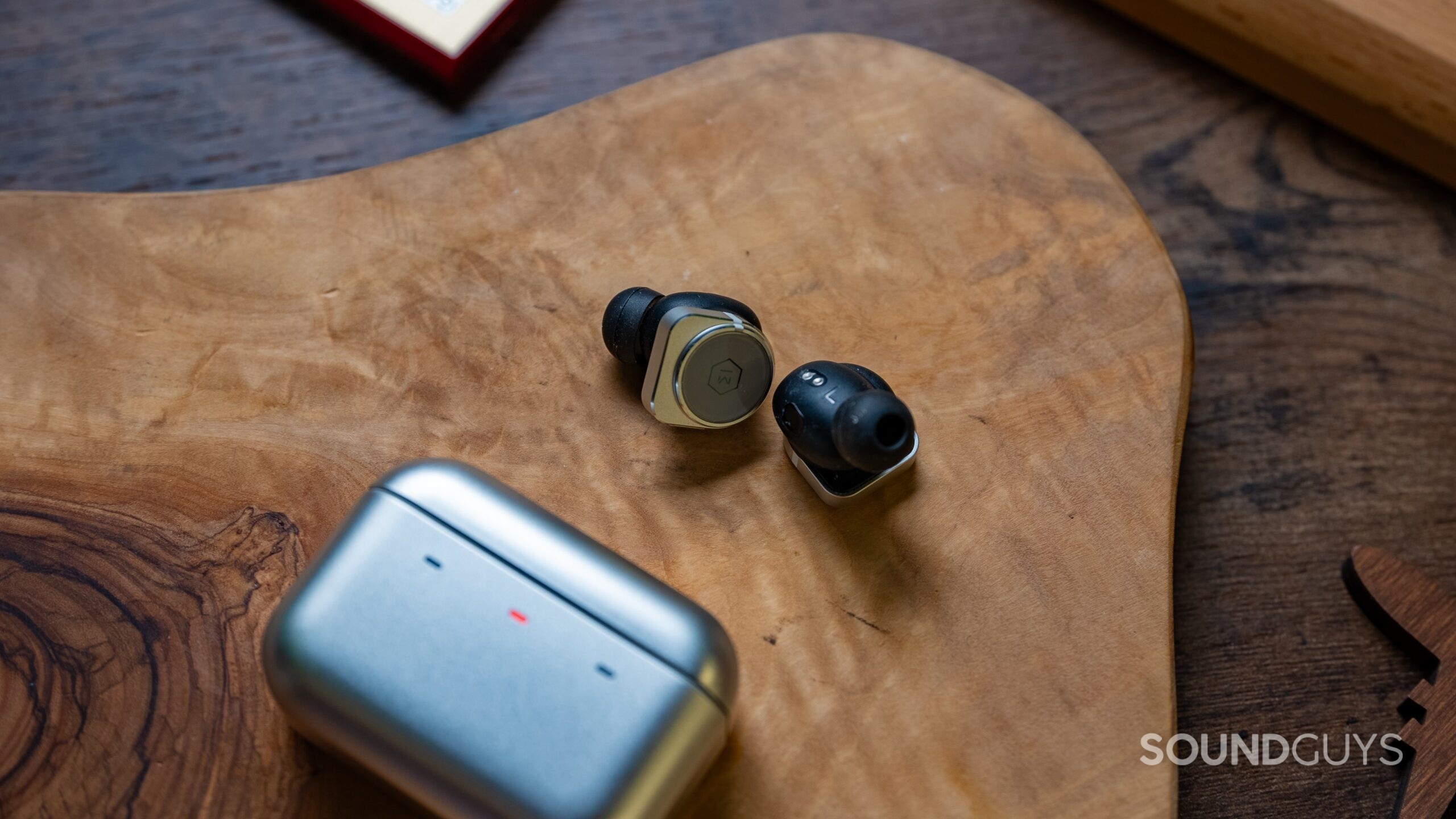 Master &amp; Dynamic MW09 earbuds next to their charging case with an LED light on the charging case with the color red.