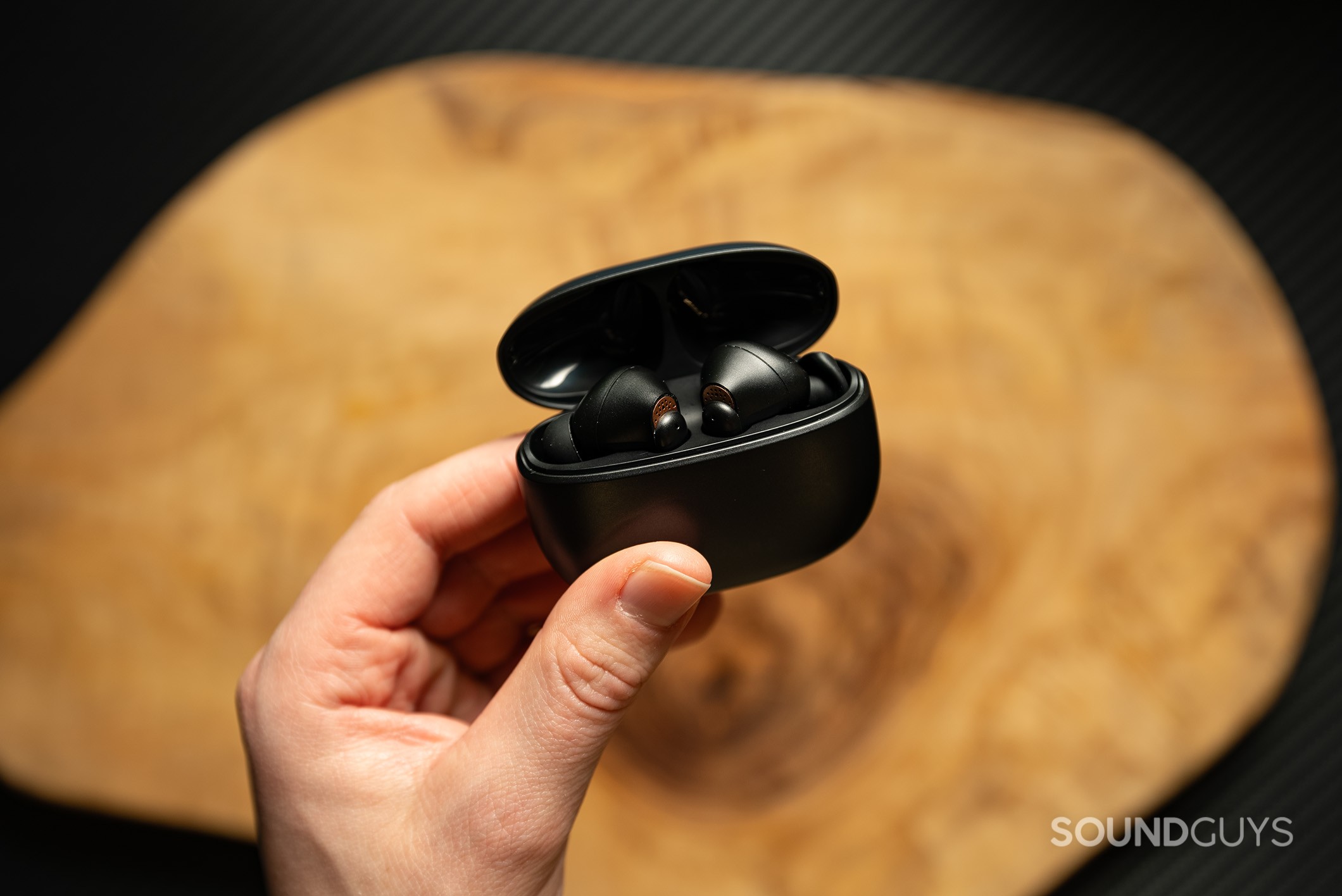 Creative Aurvana Ace earbuds in charging case held by a hand above a wood table.