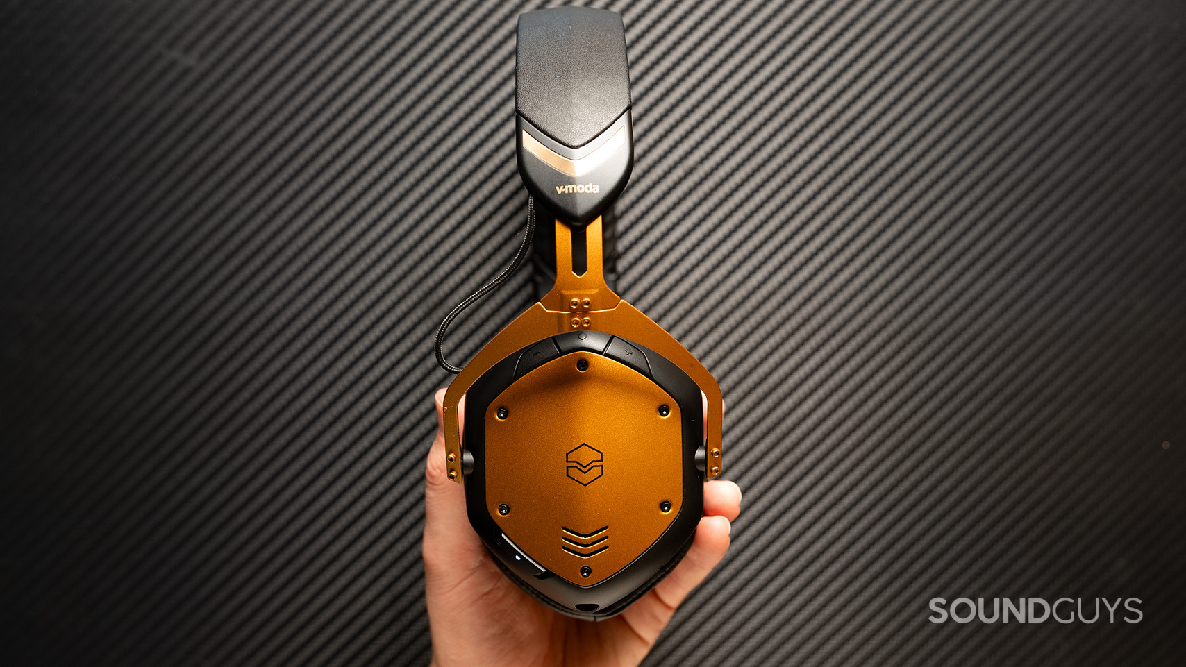 A hand holds the V-MODA Crossfade 3 Wireless with the buttons facing out. 
