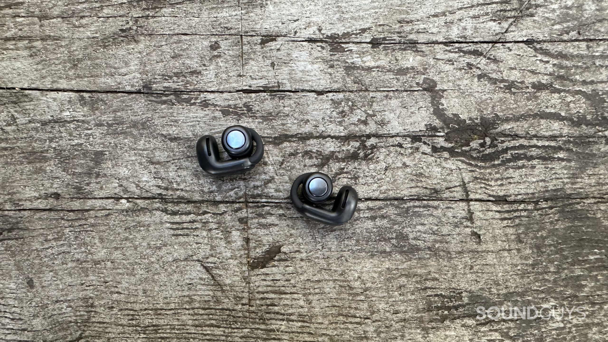 A pair of Bose Ultimate Open Earbuds with control buttons facing up on a grey bench