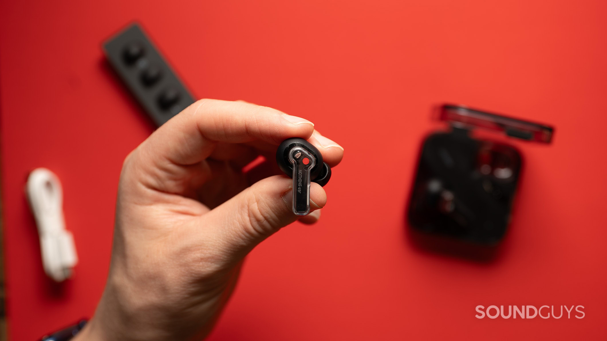 A hand holds a Nothing Ear earbud over a table with the charging case, extra ear tips, and charging cable.