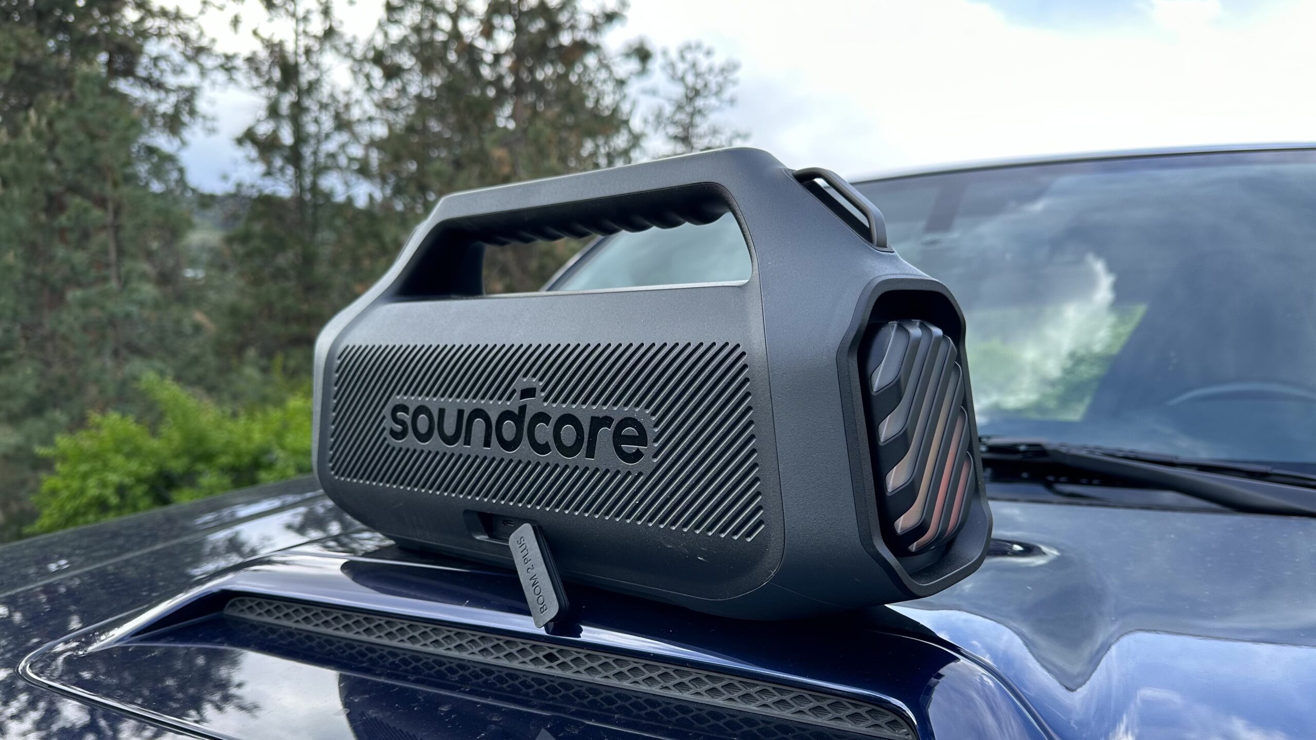 Anker Soundcore Boom 2 Plus 9 An Anker Soundcore Boom 2 Plus placed on top of an automobile hood, backside with open USB cover facing the camera.