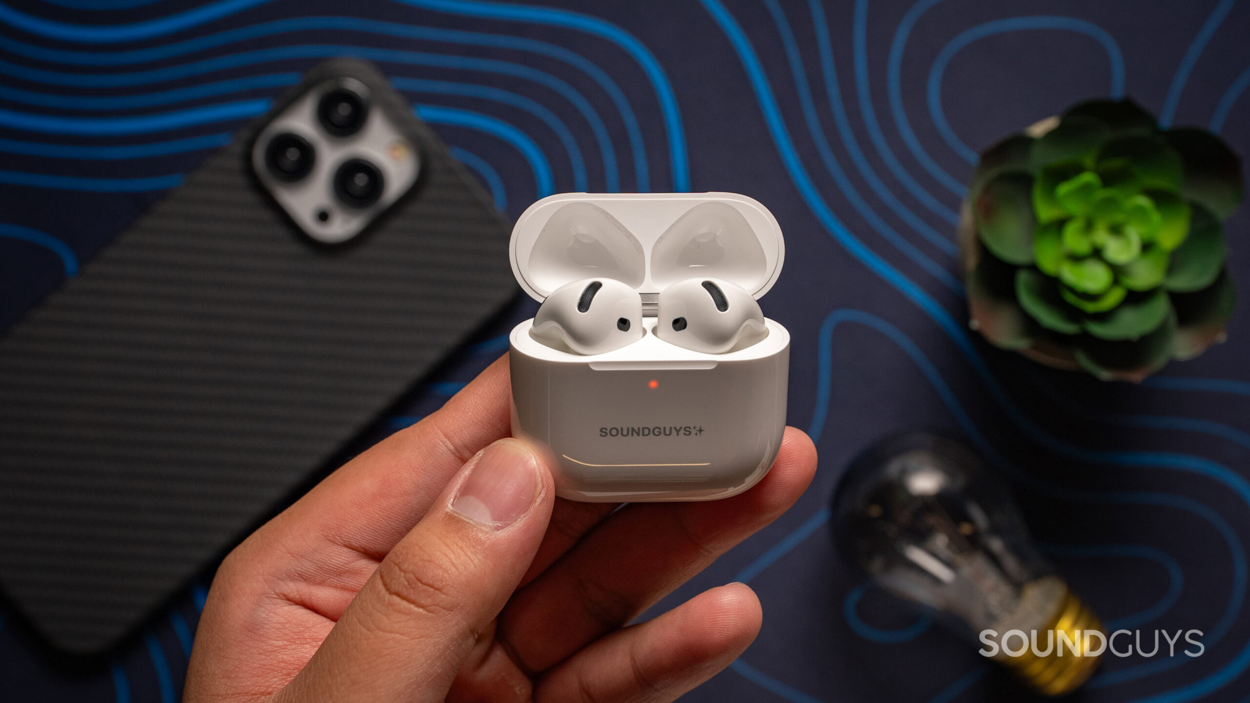 Apple AirPods 4 with Active Noise Cancellation SoundGuys engraved charging case in hand A hand holding the AirPods with Active Noise Cancellation. The earbuds are inside the charging case, and the lid is open.