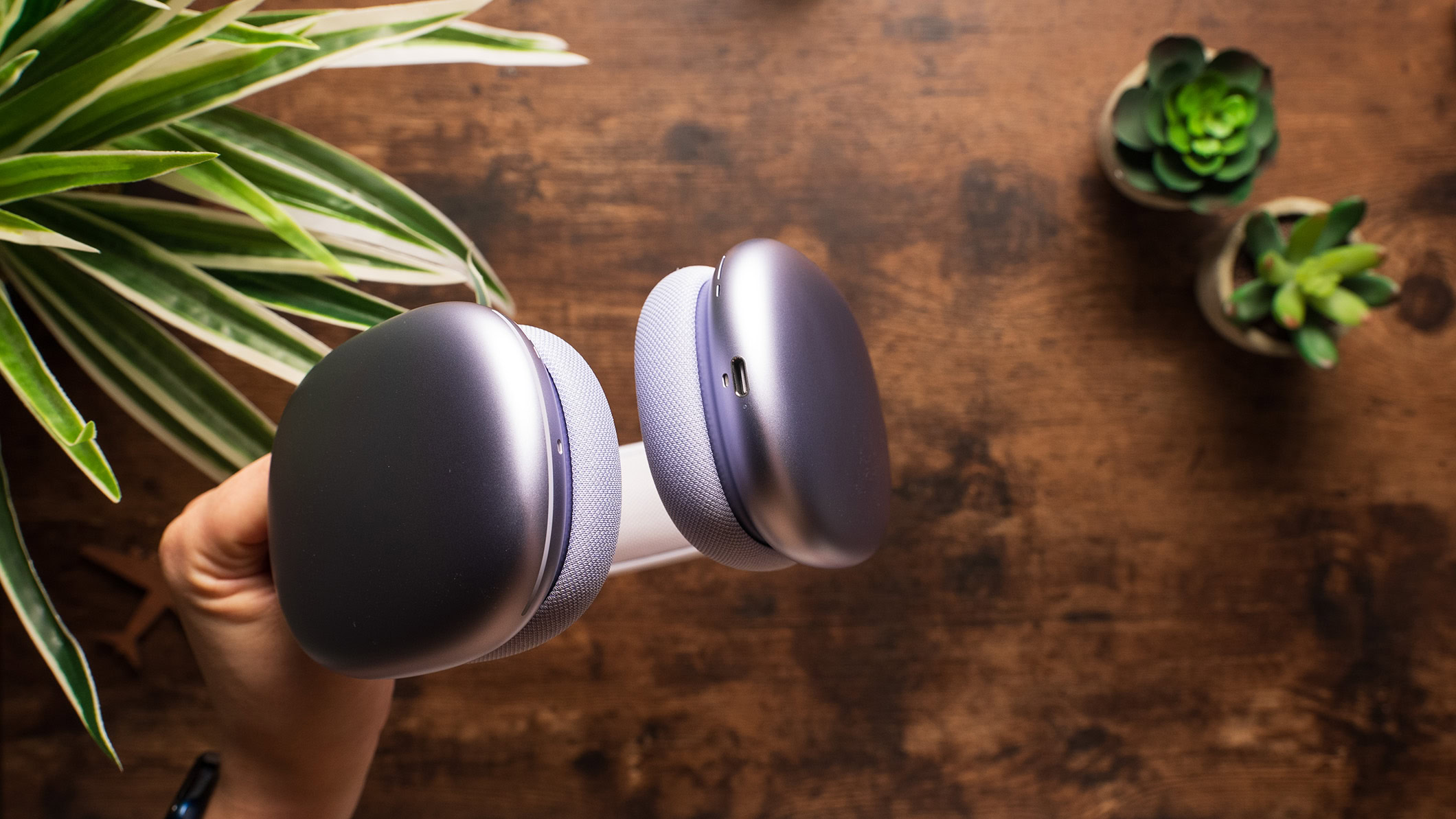 Apple AirPods Max USB C Lilac held in hand A photo of the Apple AirPods Max held in a hand above a wooden desk.