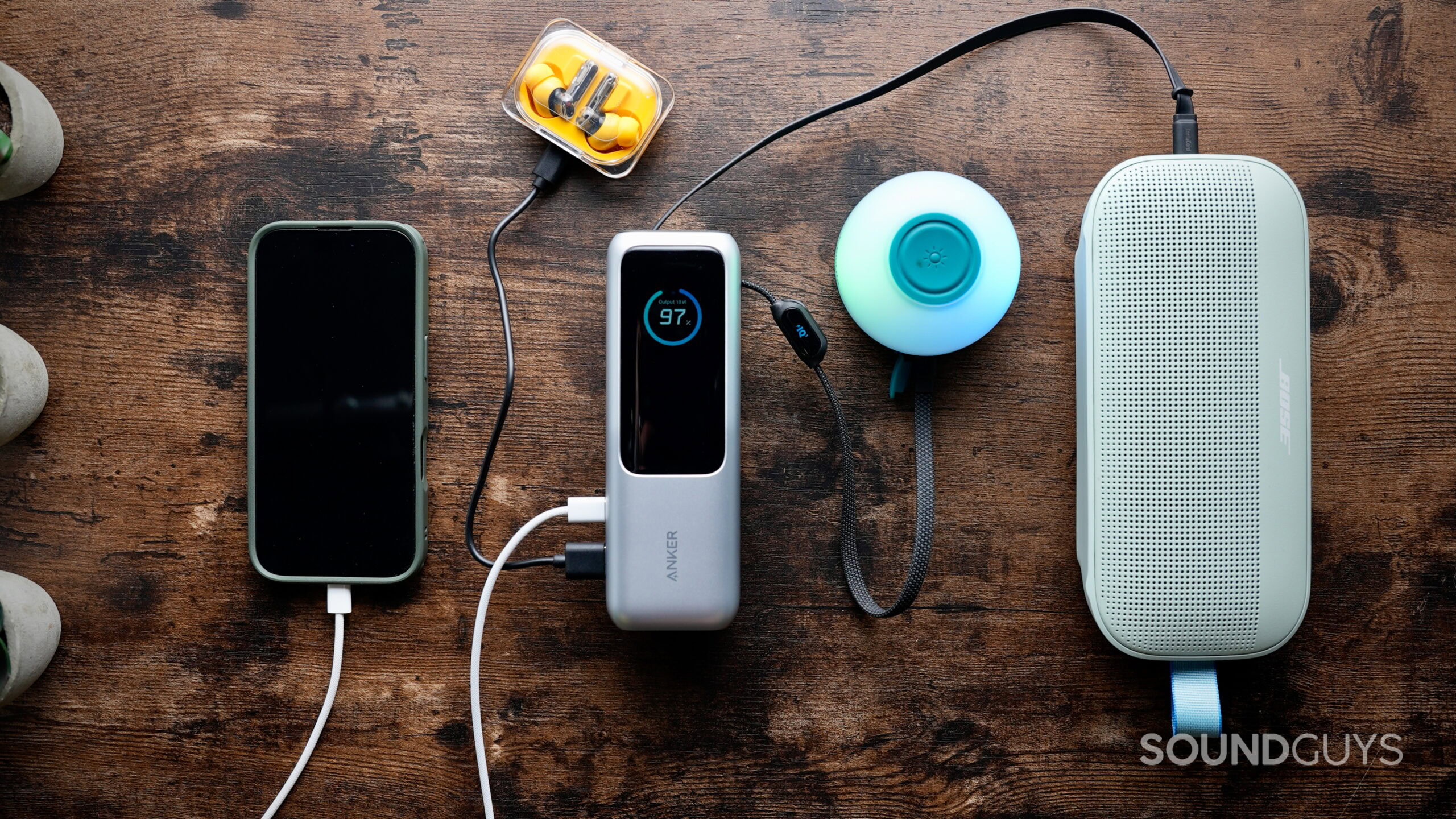 BT speaker charge out - 1 A smartphone, wireless earbuds in a yellow case, a portable power bank, a glowing portable light, and a Bose Bluetooth speaker sit on a wooden table, all connected and charging.