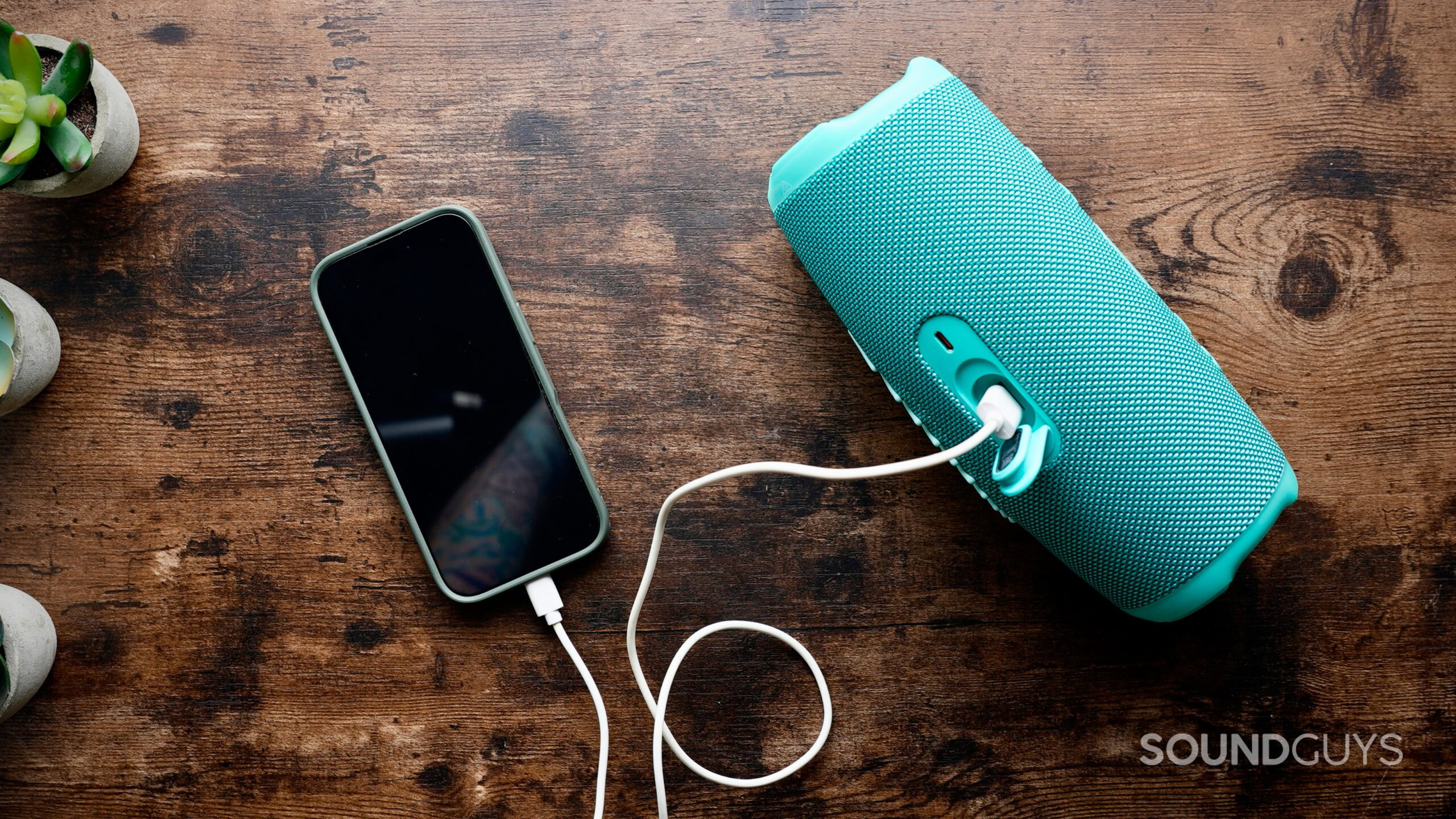 BT speaker charge out - 2 A smartphone charges from the USB output port of a teal JBL Bluetooth speaker on a wooden table, with the charging cable plugged directly into the speaker.