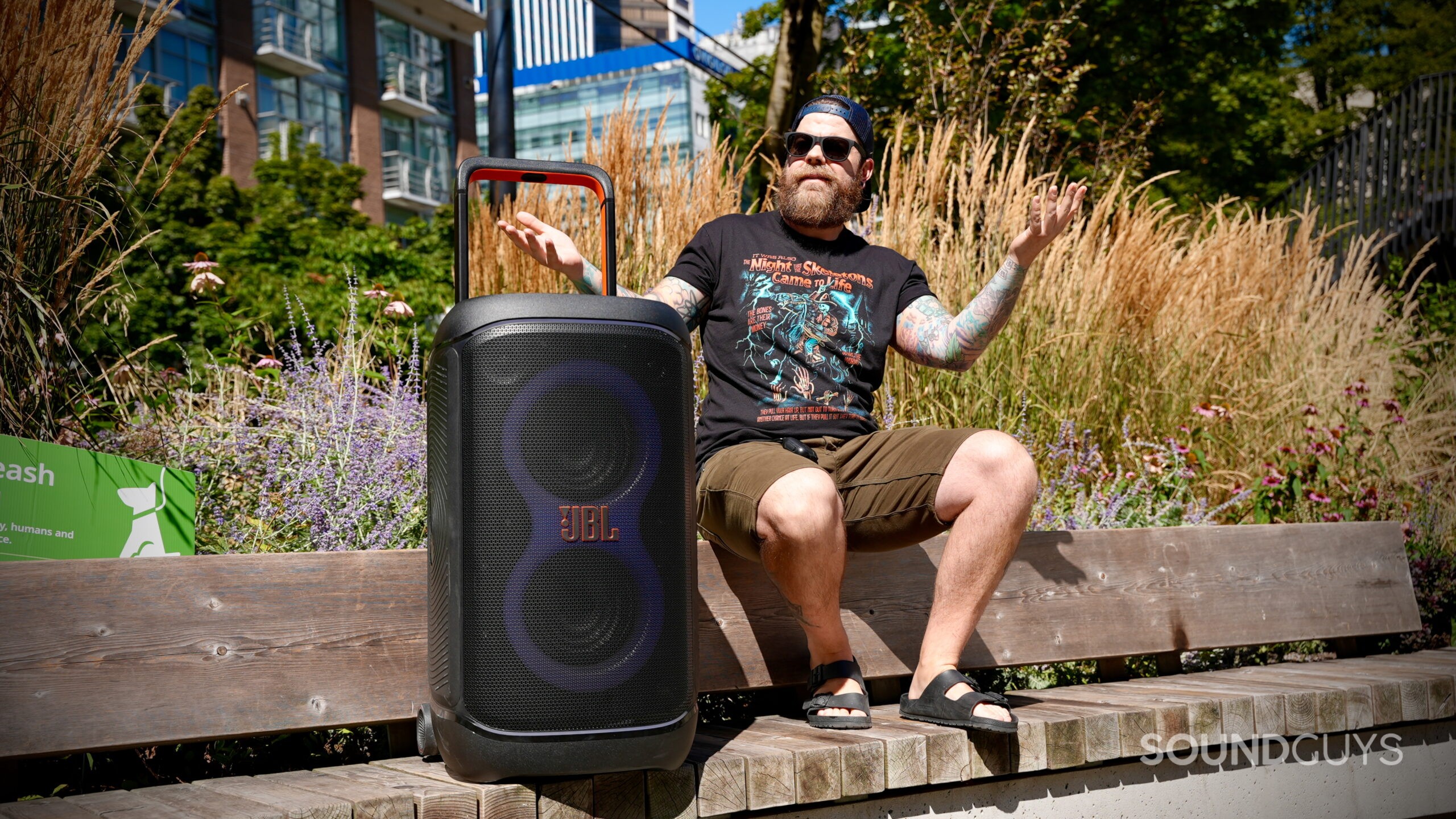 BT speakers in public - 1 A man shrugs next to a large JBL Bluetooth speaker on a park bench, reacting to loud outdoor music.