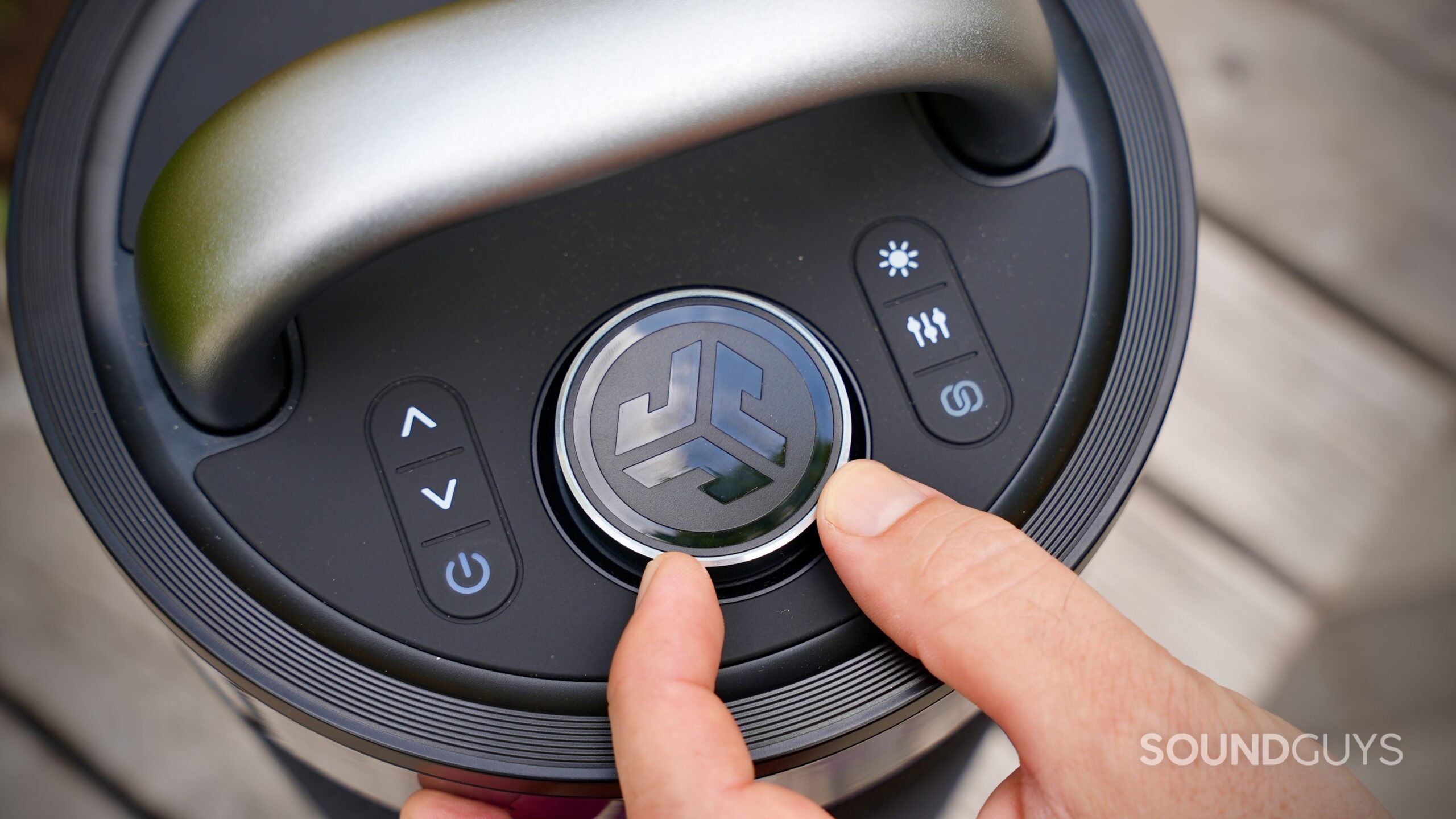 Close-up of a person pressing the central button on the top panel of the JLab Epic Party speaker. The controls include volume, power, lighting, sound modes, and stereo pairing, with a large metallic handle arching over the top.