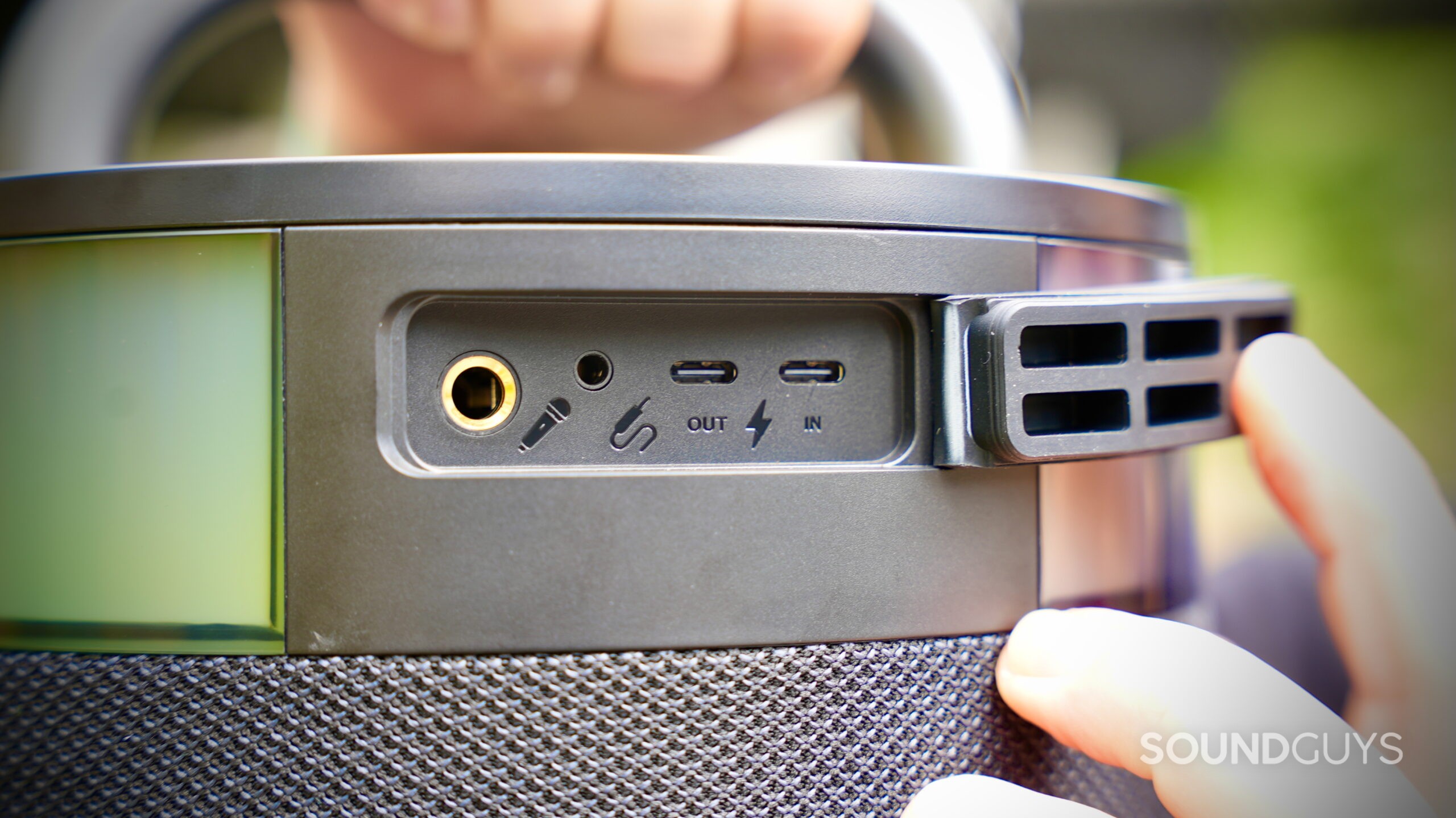 Close-up of the JLab Epic Party speaker’s rear port panel, showing inputs and outputs including a 6.35mm microphone jack, 3.5mm aux input, USB-C output, and USB-C charging input. A hand holds open the protective rubber flap covering the ports.