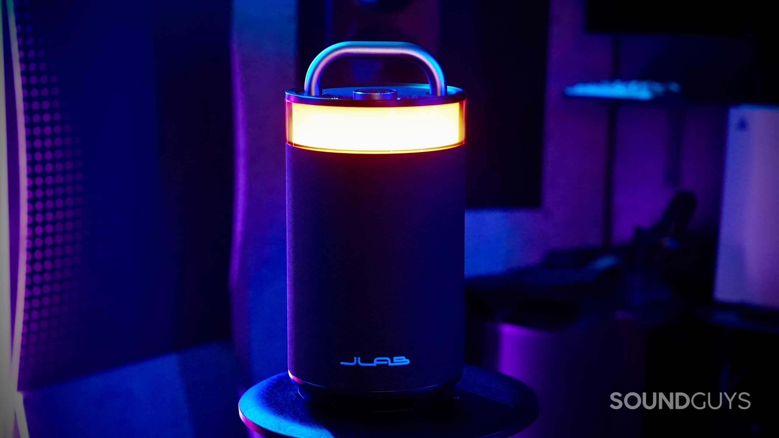 The JLab Epic Party speaker on a black stand, illuminated by vibrant blue and purple ambient lighting, with a glowing orange LED ring near the top and a metal handle above.