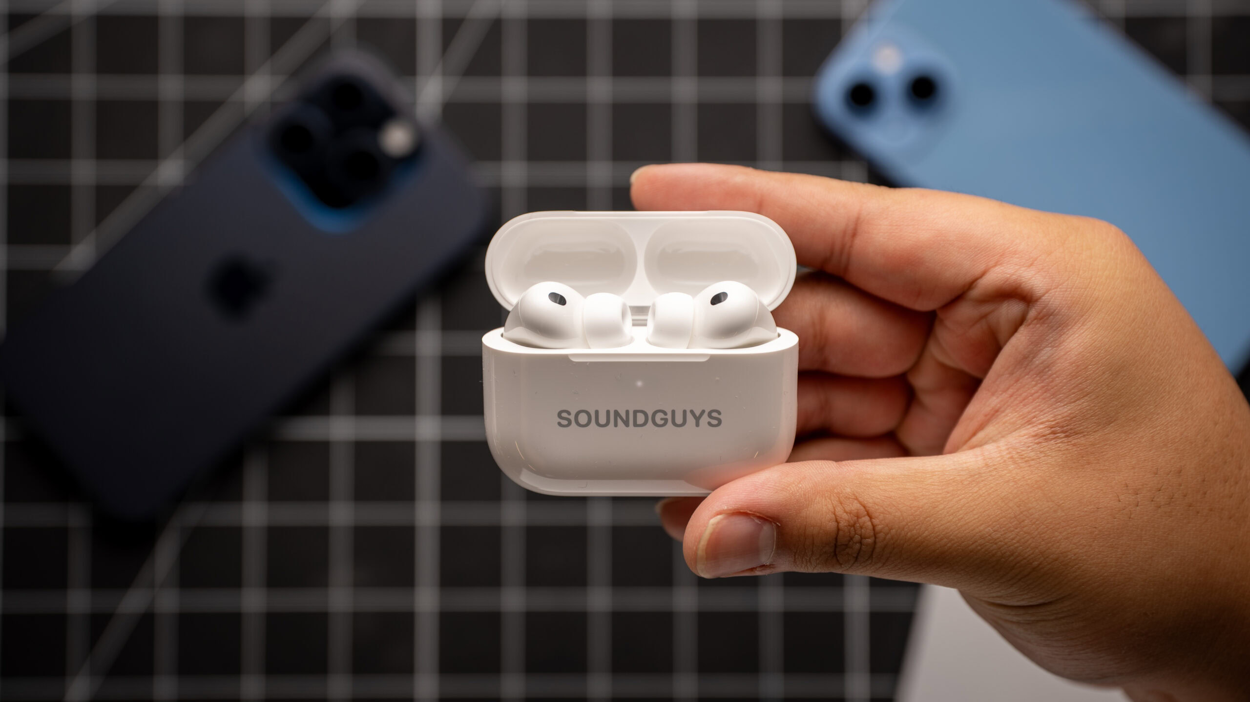 A hand holds the Apple AirPods Pro 3 in the charging case, with iPhones in the background.
