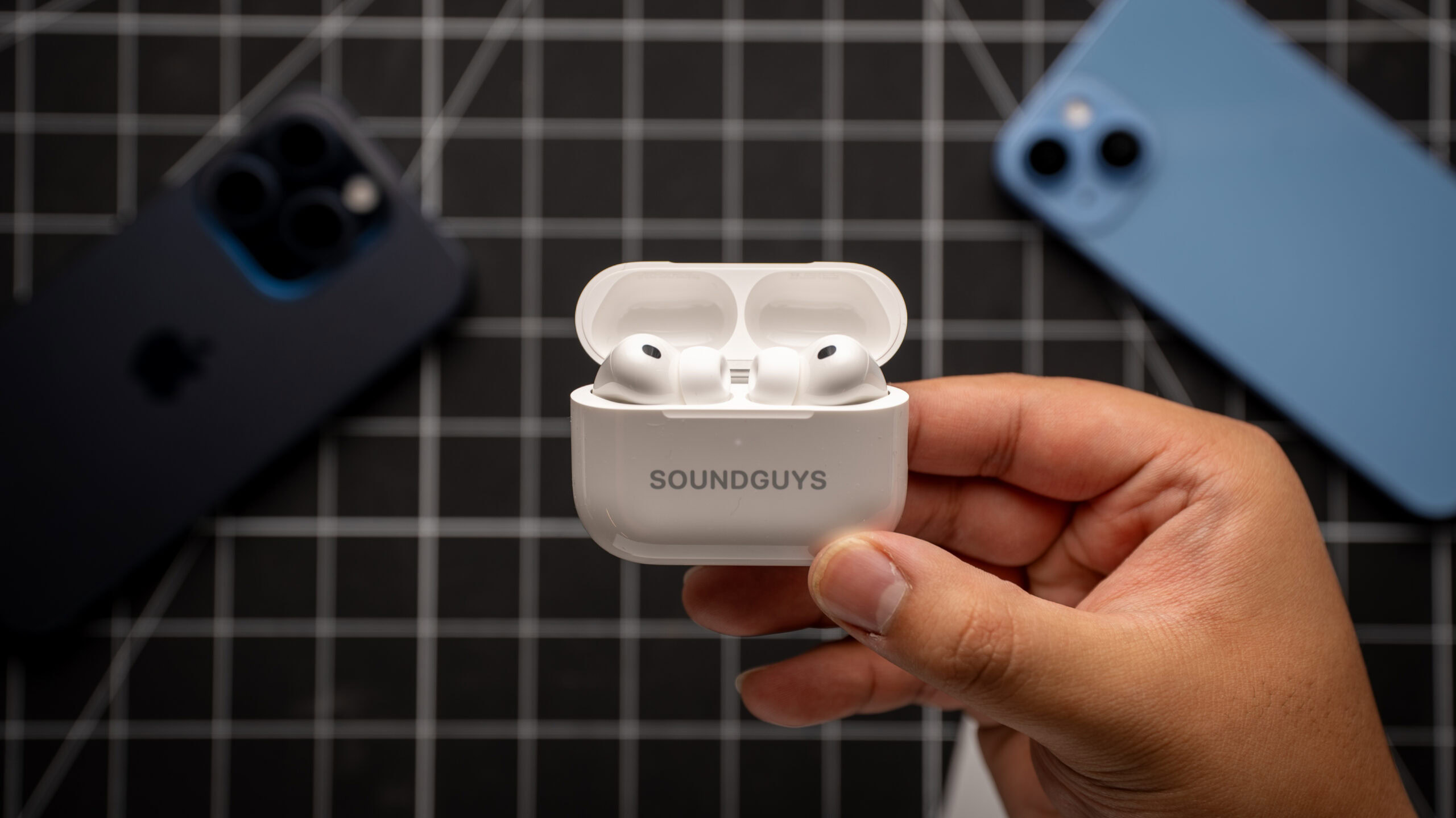 AirPods Pro 3 earbuds in charging case in hand over a table with white grid lines