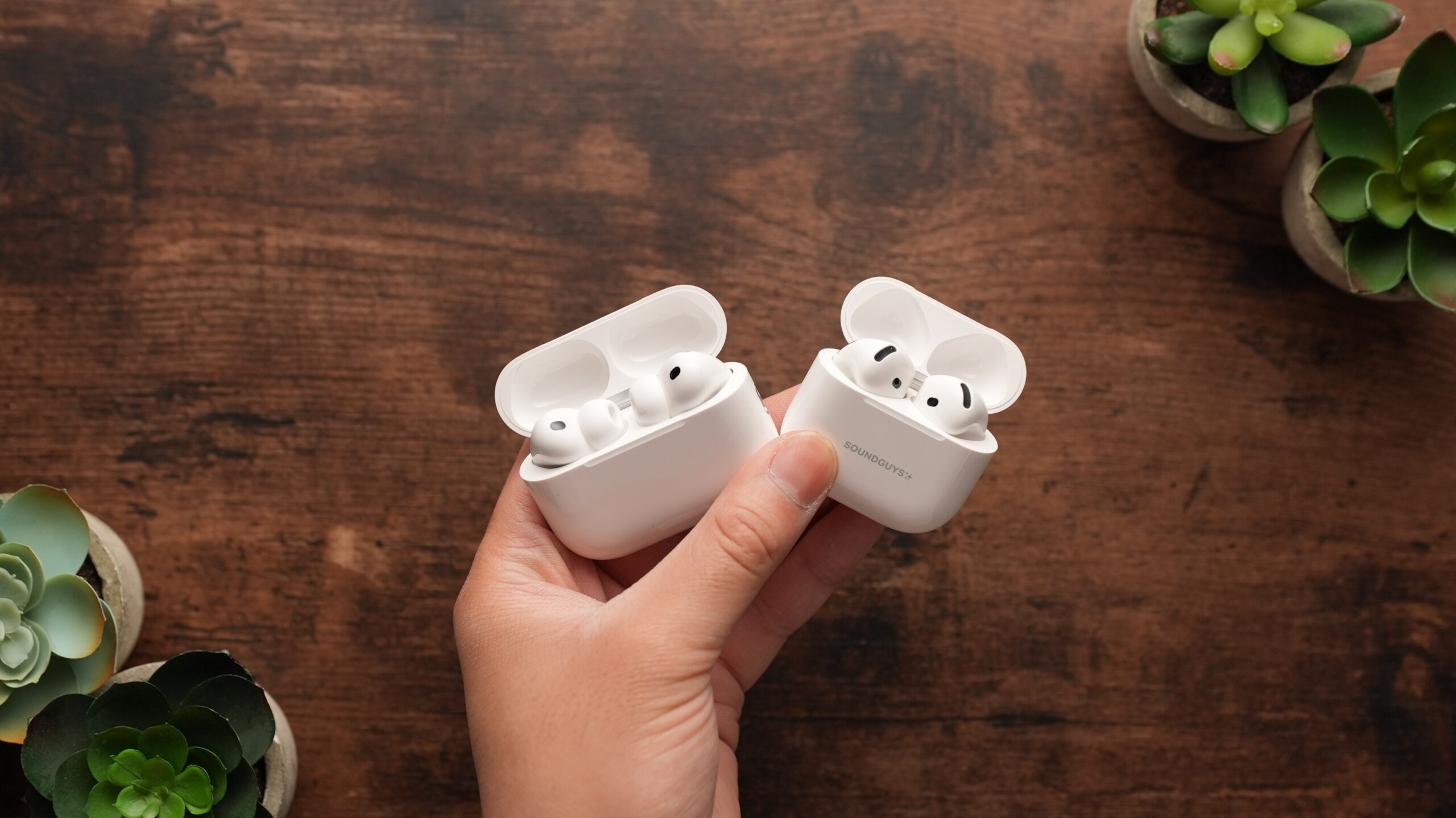 A hand holds both the AirPods Pro 3 and AirPods 4 over a wooden background.