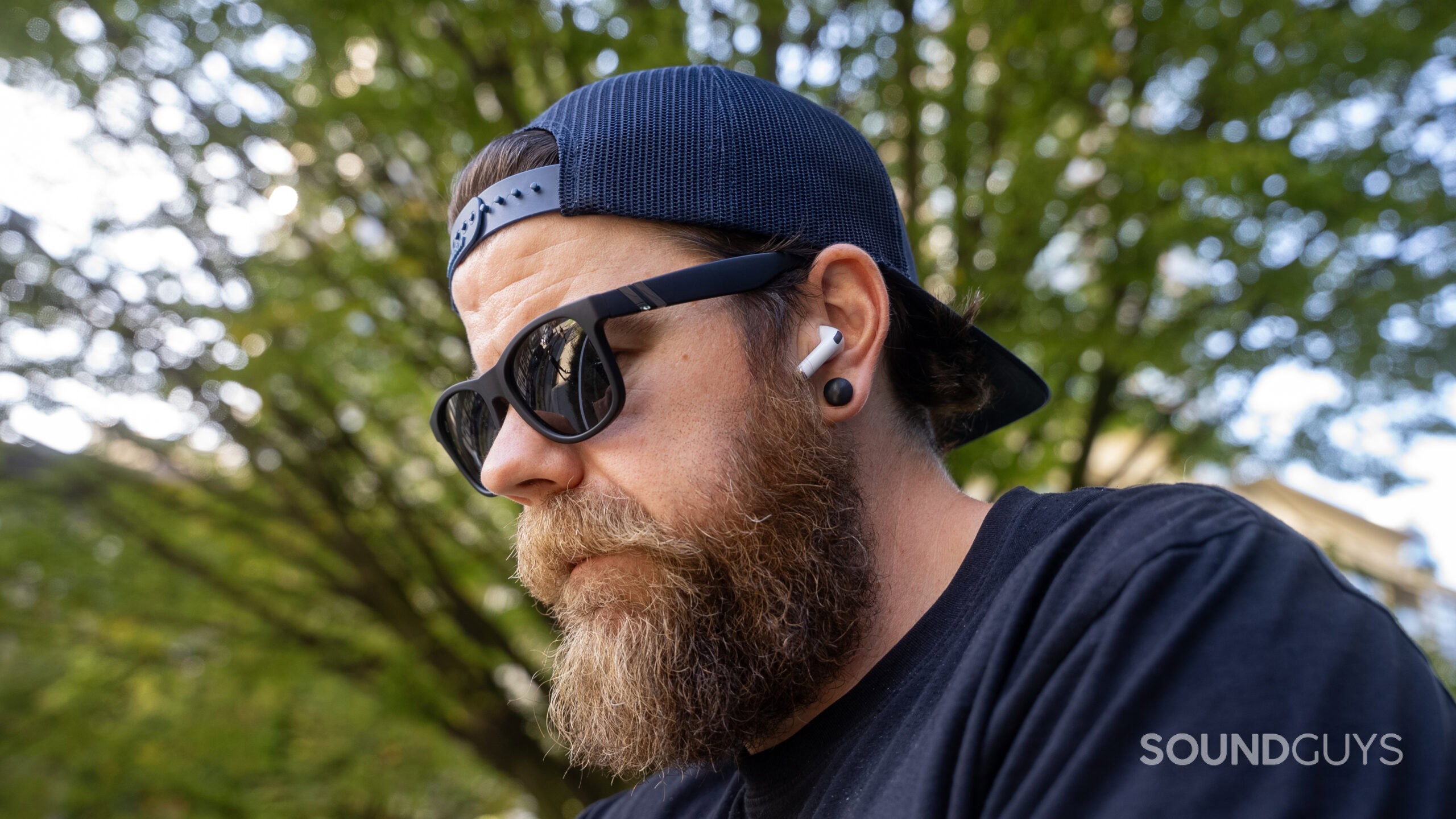 A bearded man with sunglasses wears the Apple AirPods Pro 3 outdoors.