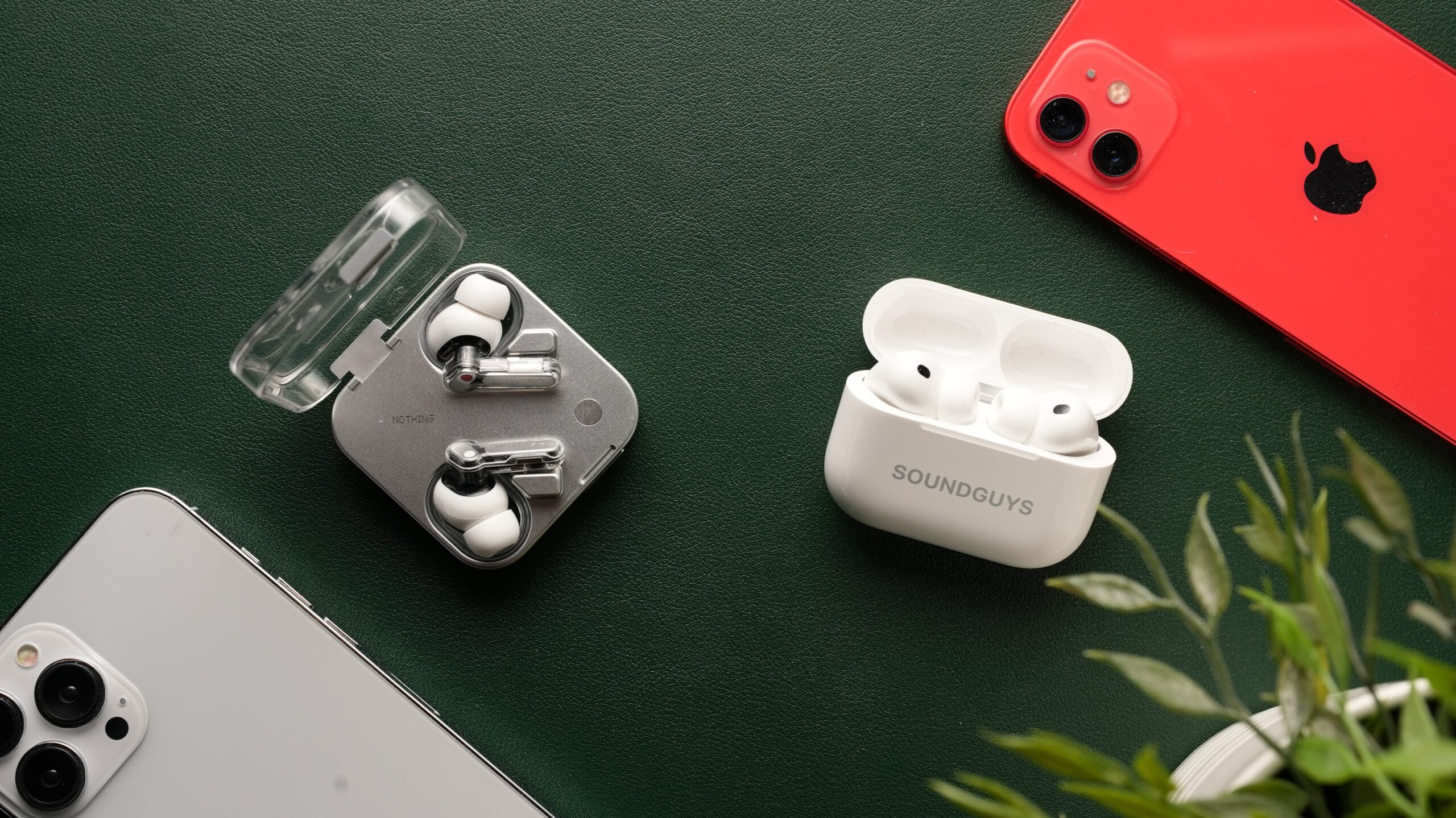 airpods-pro-3-vs-nothing-ear-3-1 The AirPods Pro 3 and the Nothing Ear (3) sit side-by-side against a dark green backdrop, surrounded by iPhones and a plant.