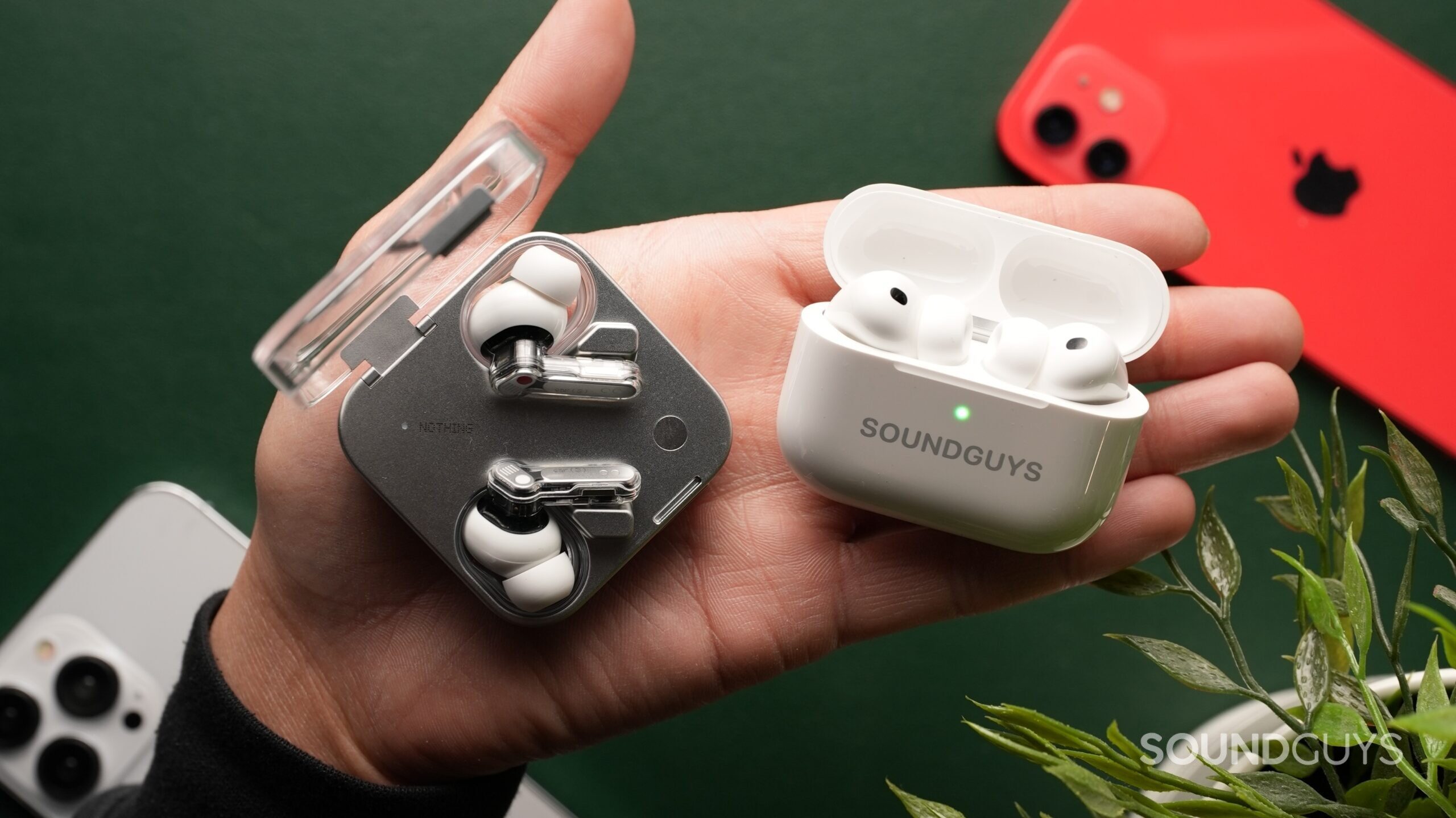 airpods-pro-3-vs-nothing-ear-3-2 The AirPods Pro 3 and the Nothing Ear (3) sit side-by-side on a palm against a dark green backdrop, surrounded by iPhones and a plant.