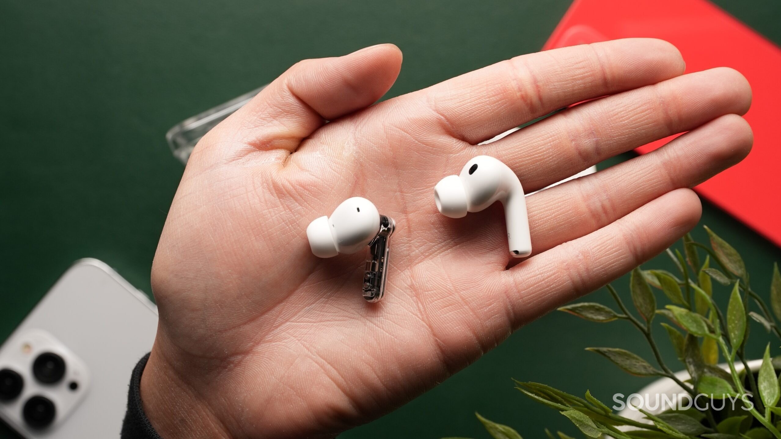 airpods-pro-3-vs-nothing-ear-3-3 The AirPods Pro 3 and the Nothing Ear (3) sit out of their cases side-by-side on a palm against a dark green backdrop, surrounded by iPhones and a plant.