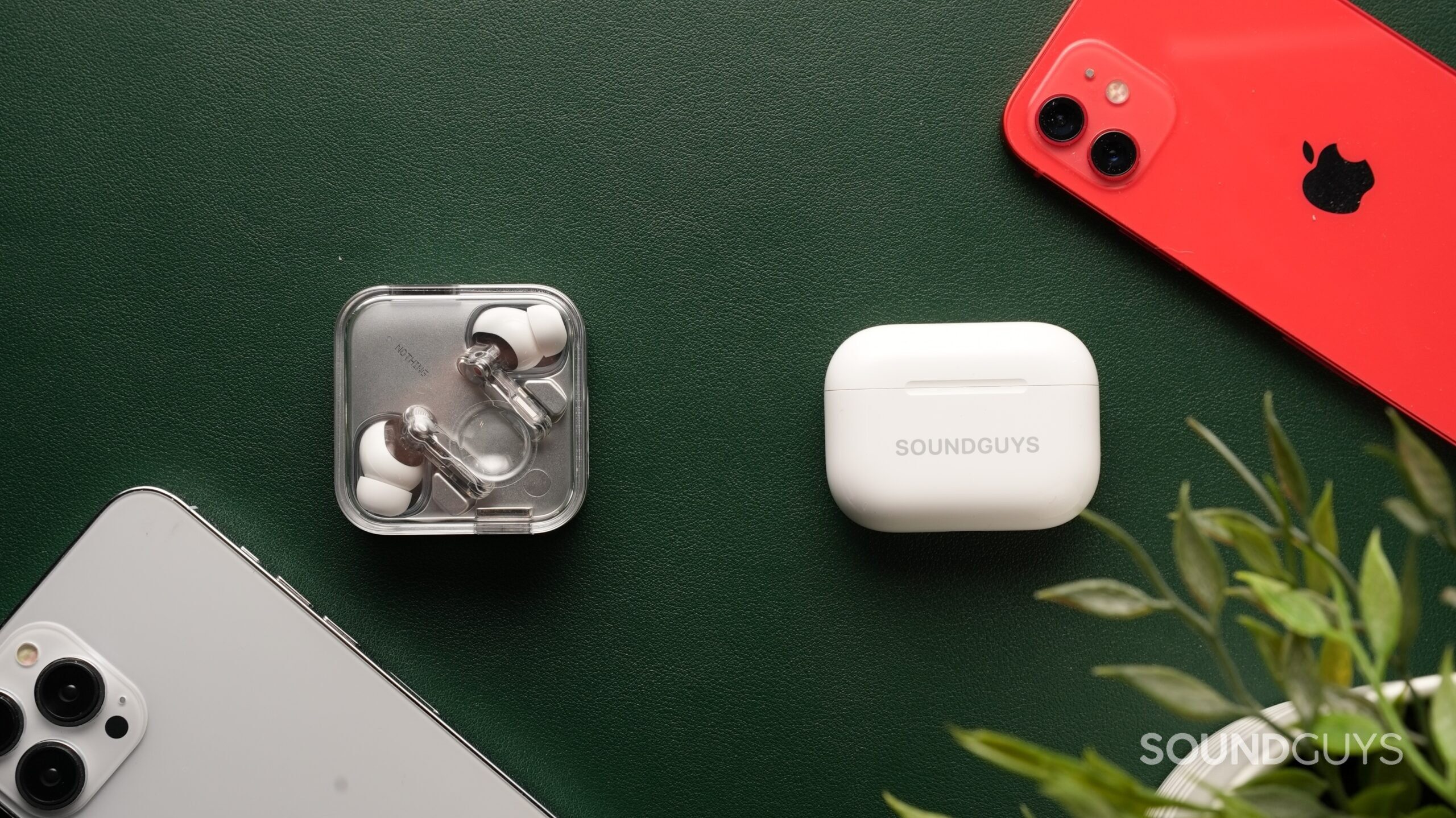airpods-pro-3-vs-nothing-ear-3-4 The AirPods Pro 3 and the Nothing Ear (3) sit side-by-side against a dark green backdrop, surrounded by iPhones and a plant.