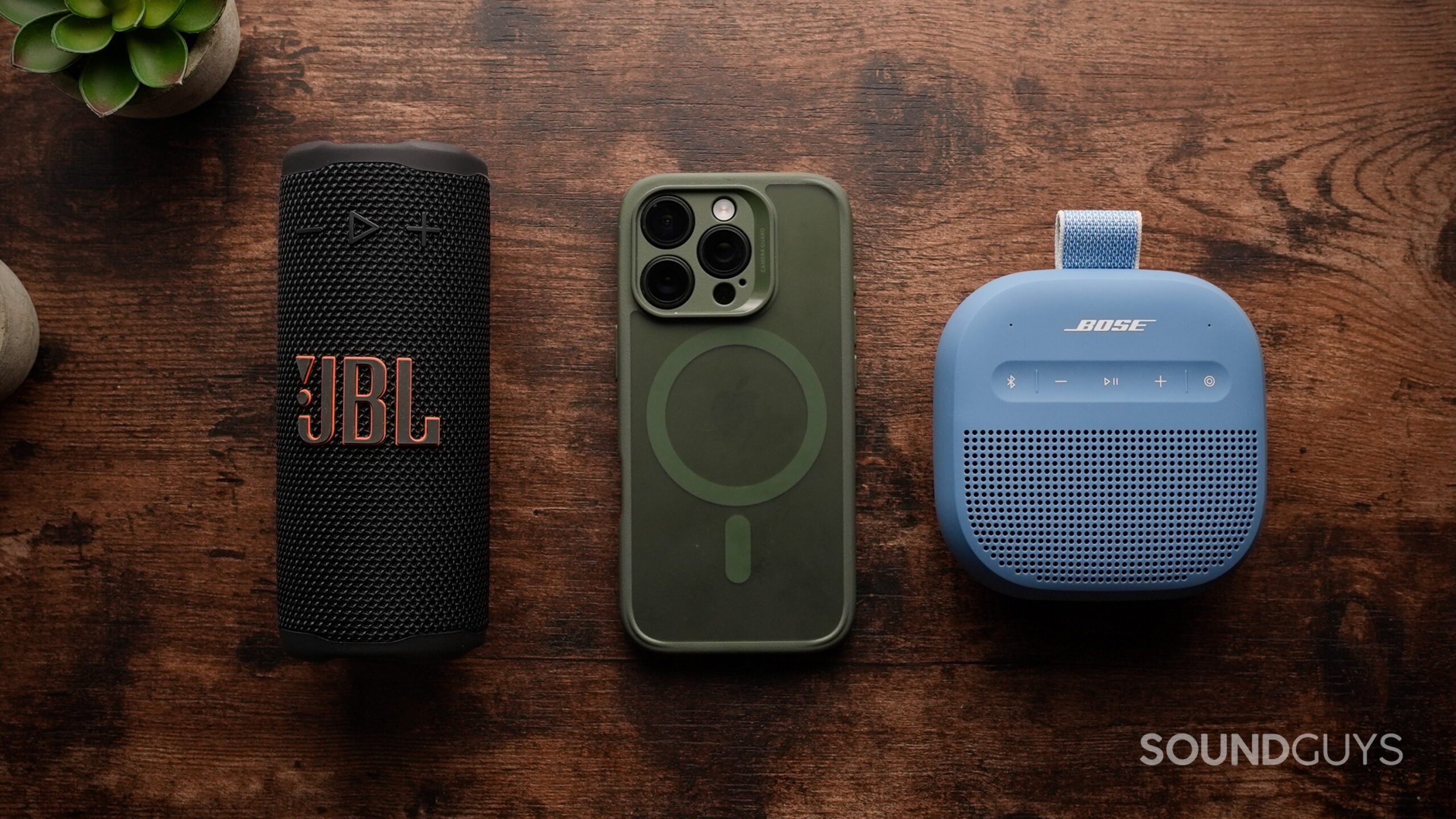 A JBL Grip, an iPhone 16 Pro, and a Bose SoundLink Micro (2nd Gen) on a wood surface.