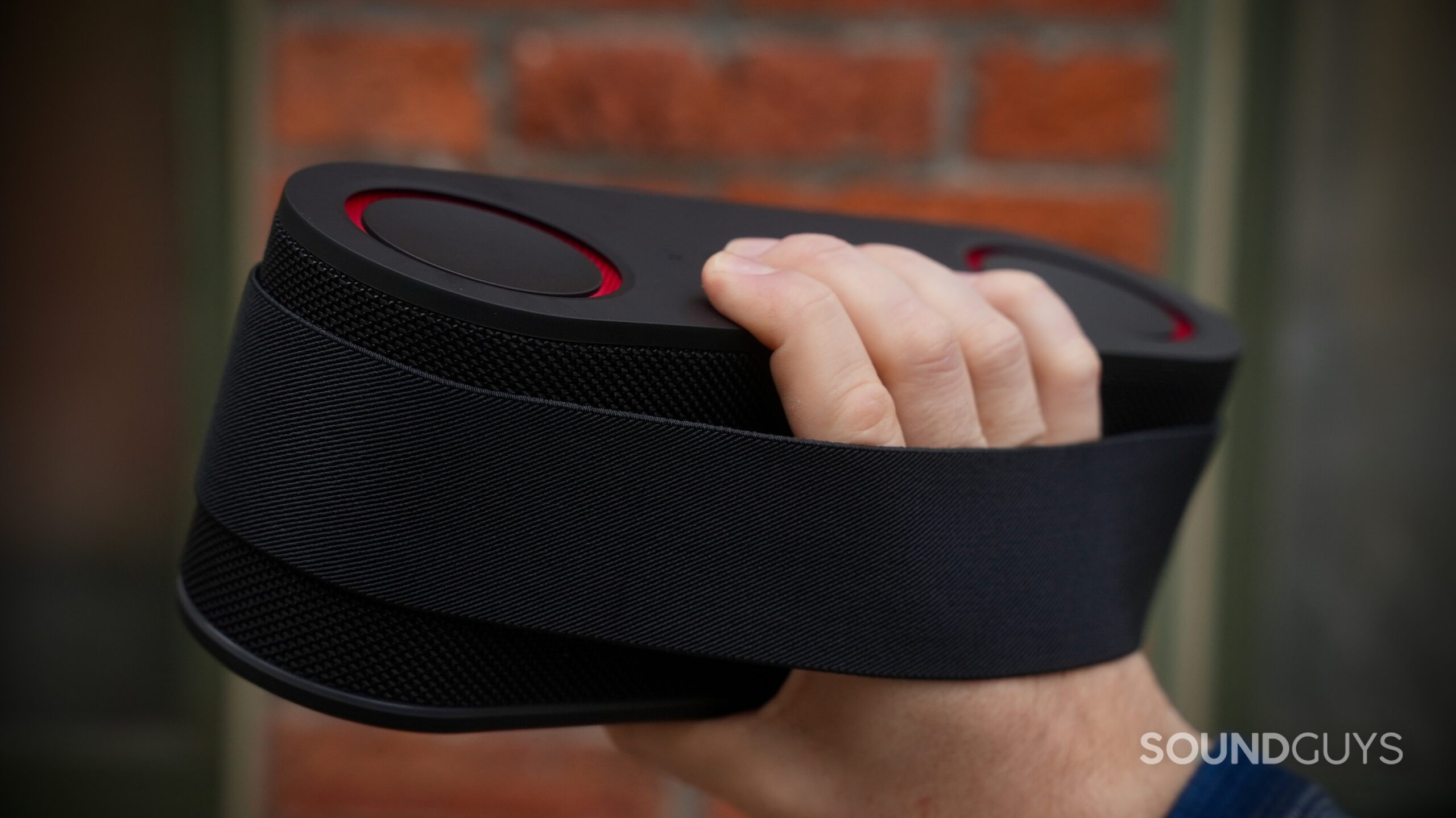A hand holding the LG xboom Bounce speaker by the back, with its attachment strap wrapped around his hand.